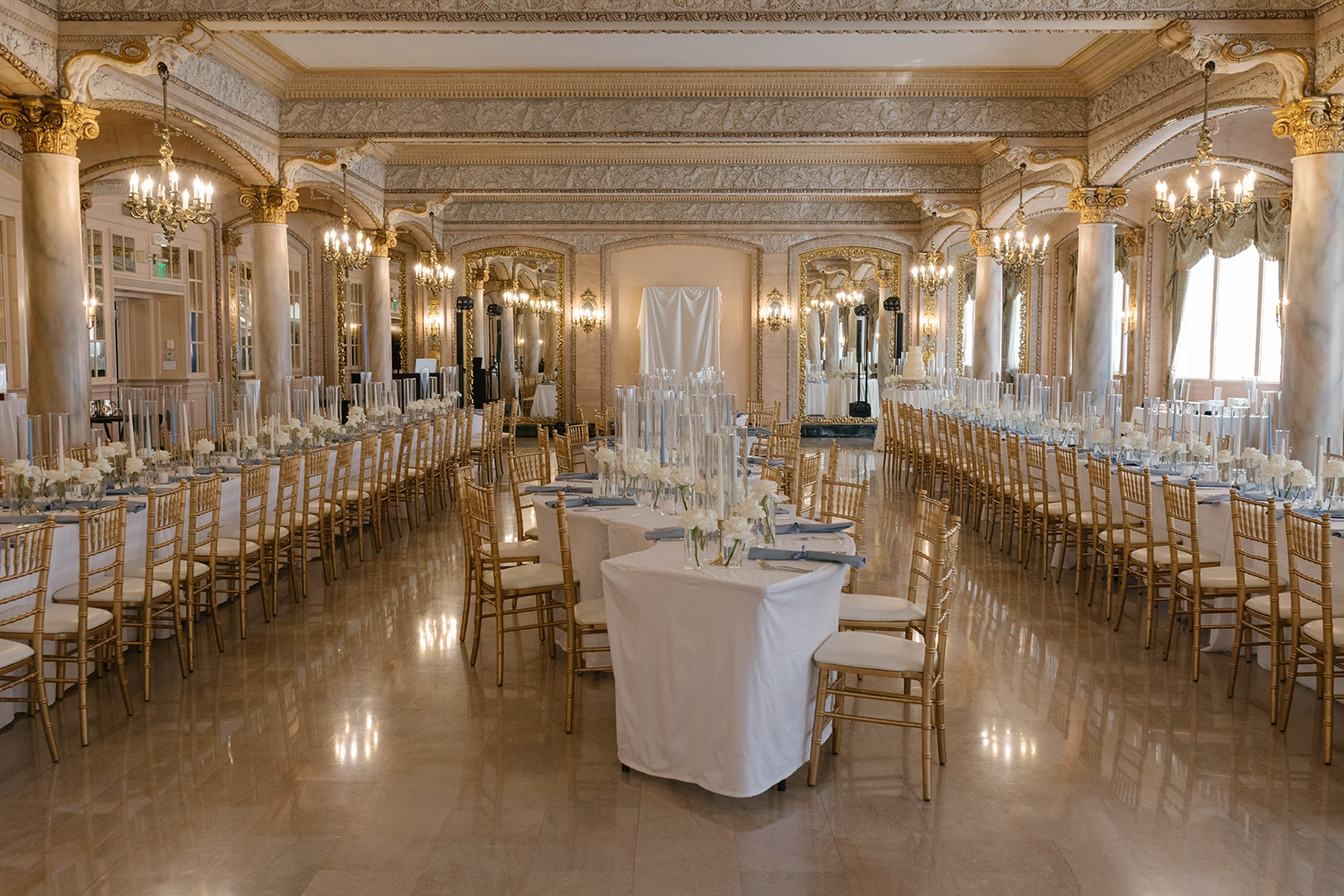 A grand, opulent ballroom prepared for a wedding reception, featuring long banquet tables covered in white linens, gold Chiavari chairs, white floral centerpieces with tall glass cylinder candles, marble floors, ornate gold-trimmed columns, and classical 