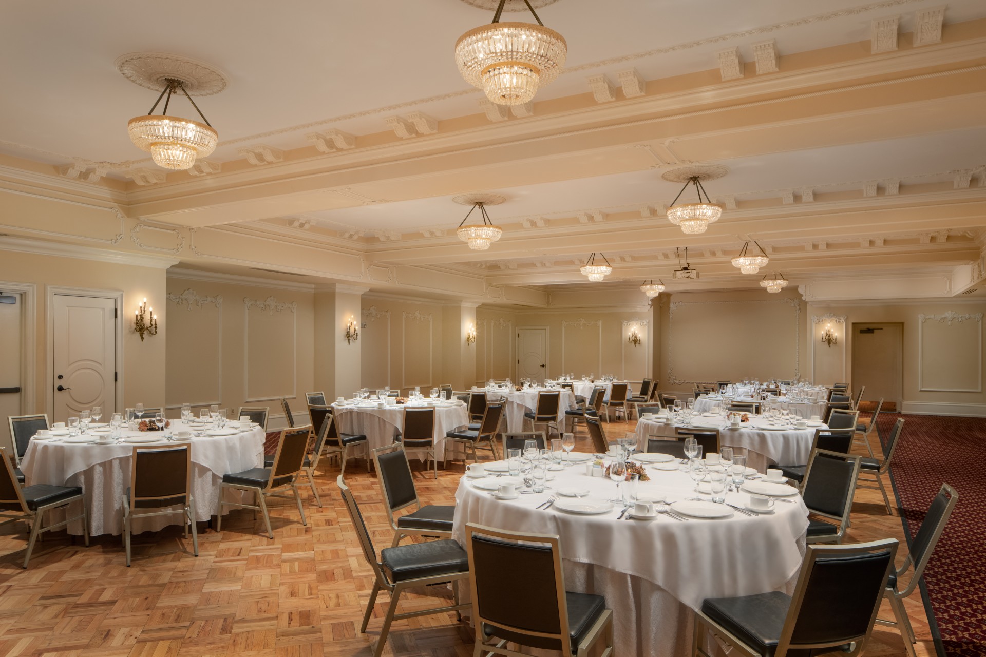 A elegant ballroom set for a banquet with round white-draped tables, black upholstered chairs, light parquet wood flooring, coffered ceilings with tier crystal bowl light fixtures, and cream walls with classic molding.
