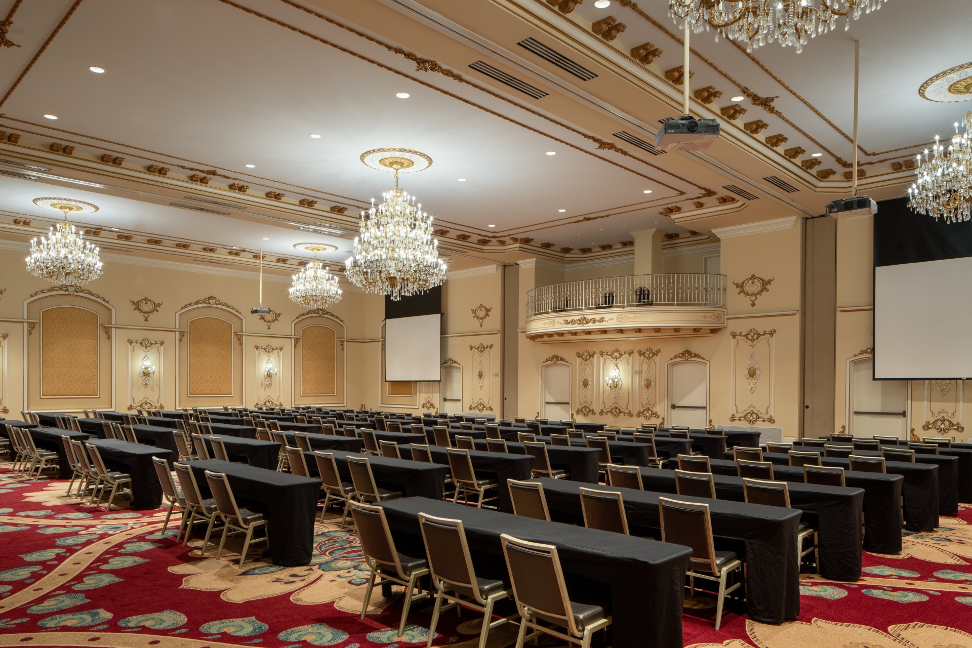 A grand, opulent ballroom configured classroom-style with black-draped tables and gold chairs, featuring crystal chandeliers hanging from an ornate gold-trimmed ceiling, projection screens, a mezzanine balcony, and vibrant red floral carpet.