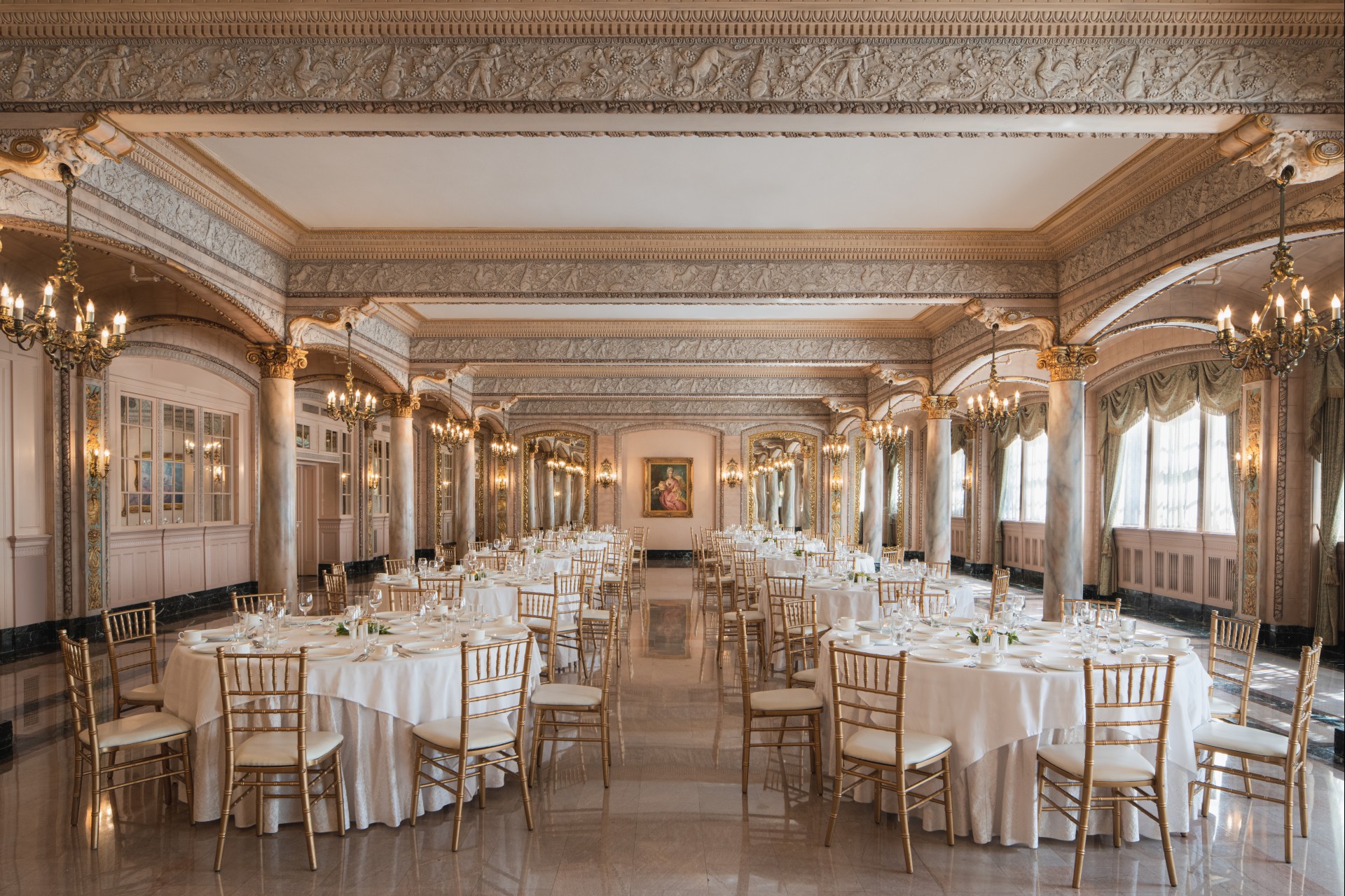 A regal banquet hall set up for a fine dining event, featuring round tables with white linens, gold Chiavari chairs, polished marble floors, classical stone arches with relief carvings, and brass chandeliers.