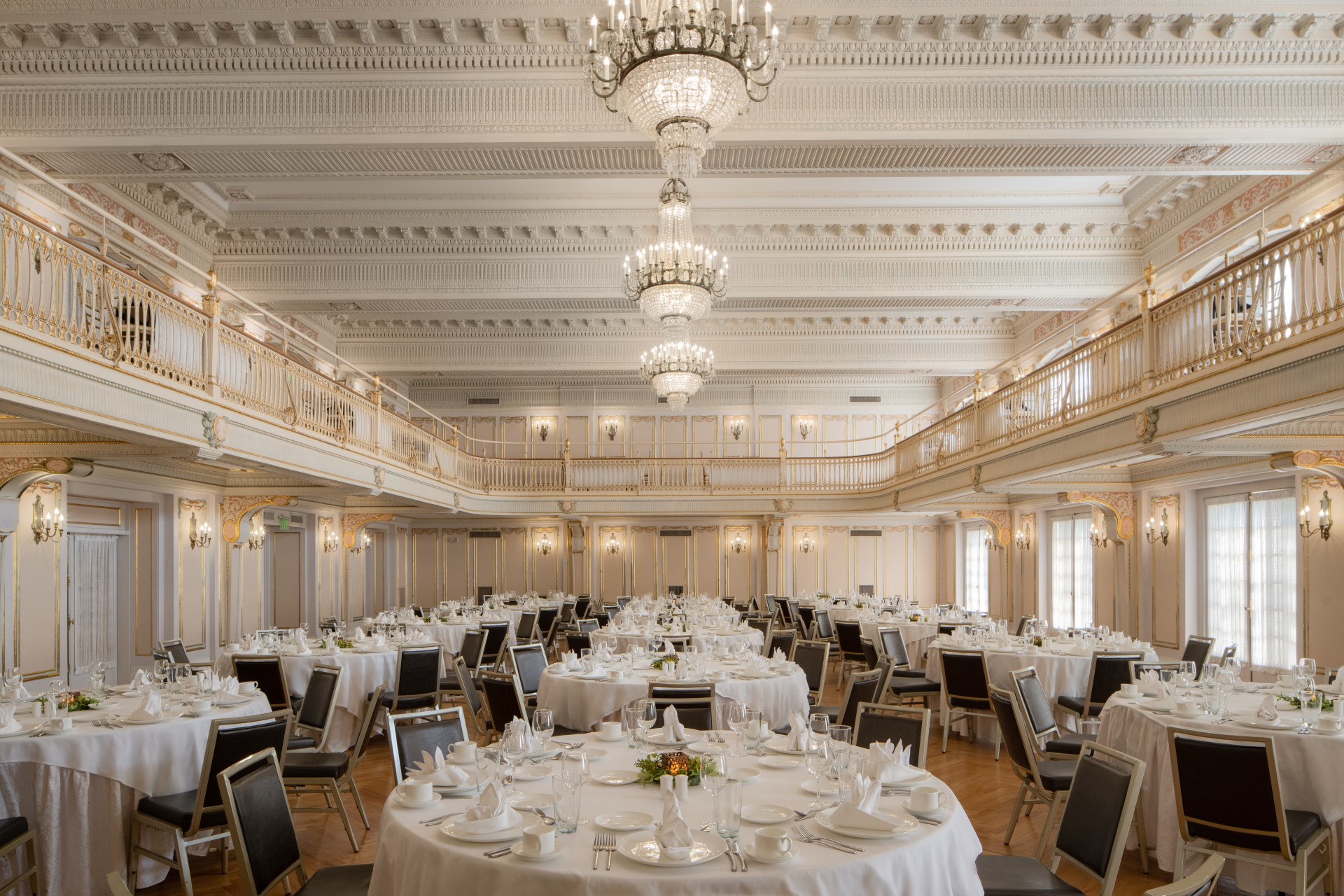 A grand, double-height ballroom set for a banquet with round tables in white linens, surrounded by a two-tier mezzanine balcony with ornate railings, elaborate crystal chandeliers, and detailed white-and-gold crown molding.