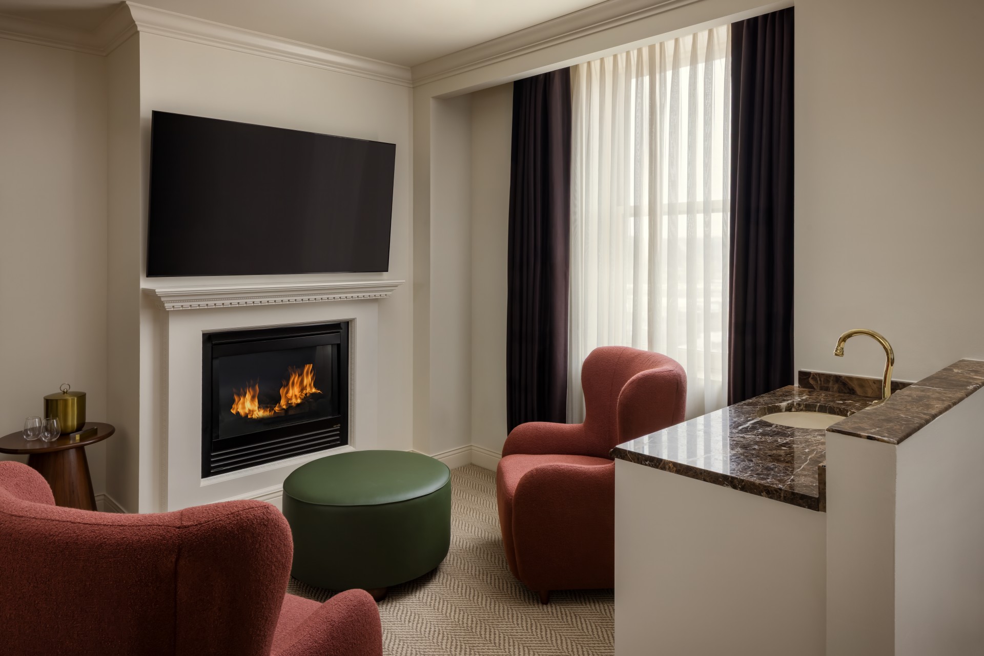 A cozy, modern hotel suite sitting area featuring a glowing gas fireplace mounted beneath a flat-screen TV, two plush dusty-rose wingback armchairs, a dark green circular ottoman, sheer white curtains flanked by dark drapes, and a marble-topped wet bar in
