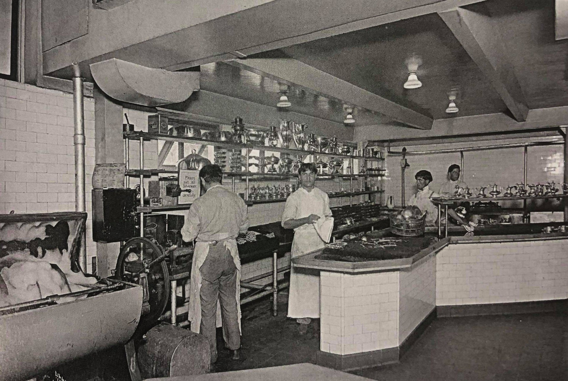 A vintage black-and-white archival photograph showing hotel kitchen staff in aprons polishing silver serveware surrounded by tiled counters, metal shelving lined with urns, and early industrial kitchen machinery.