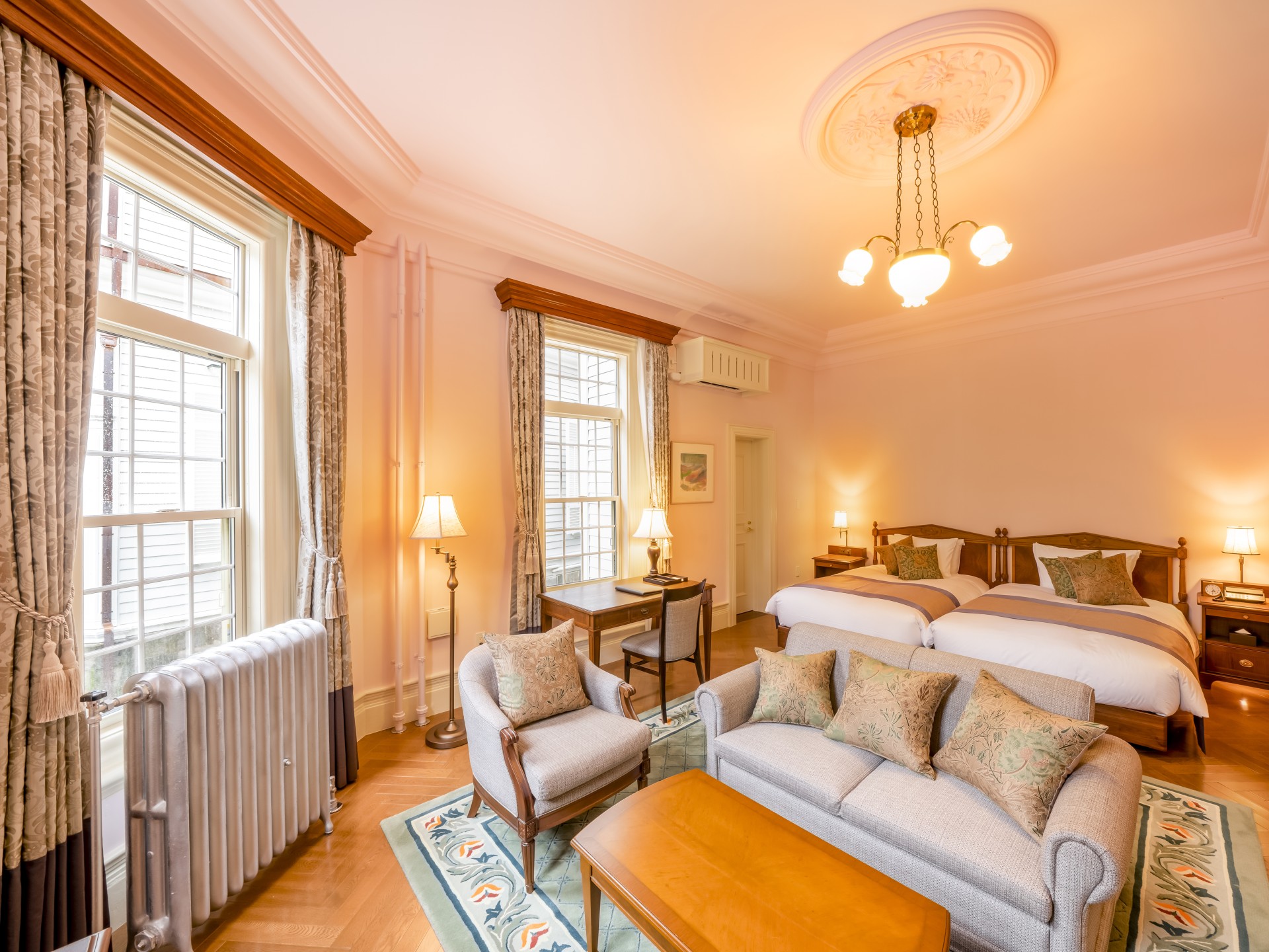 An elegant hotel suite with soft pink walls, featuring two twin beds with wooden headboards, a sitting area with a grey couch and armchair over a patterned rug, a wooden writing desk between two large windows with patterned drapes, a vintage ceiling light