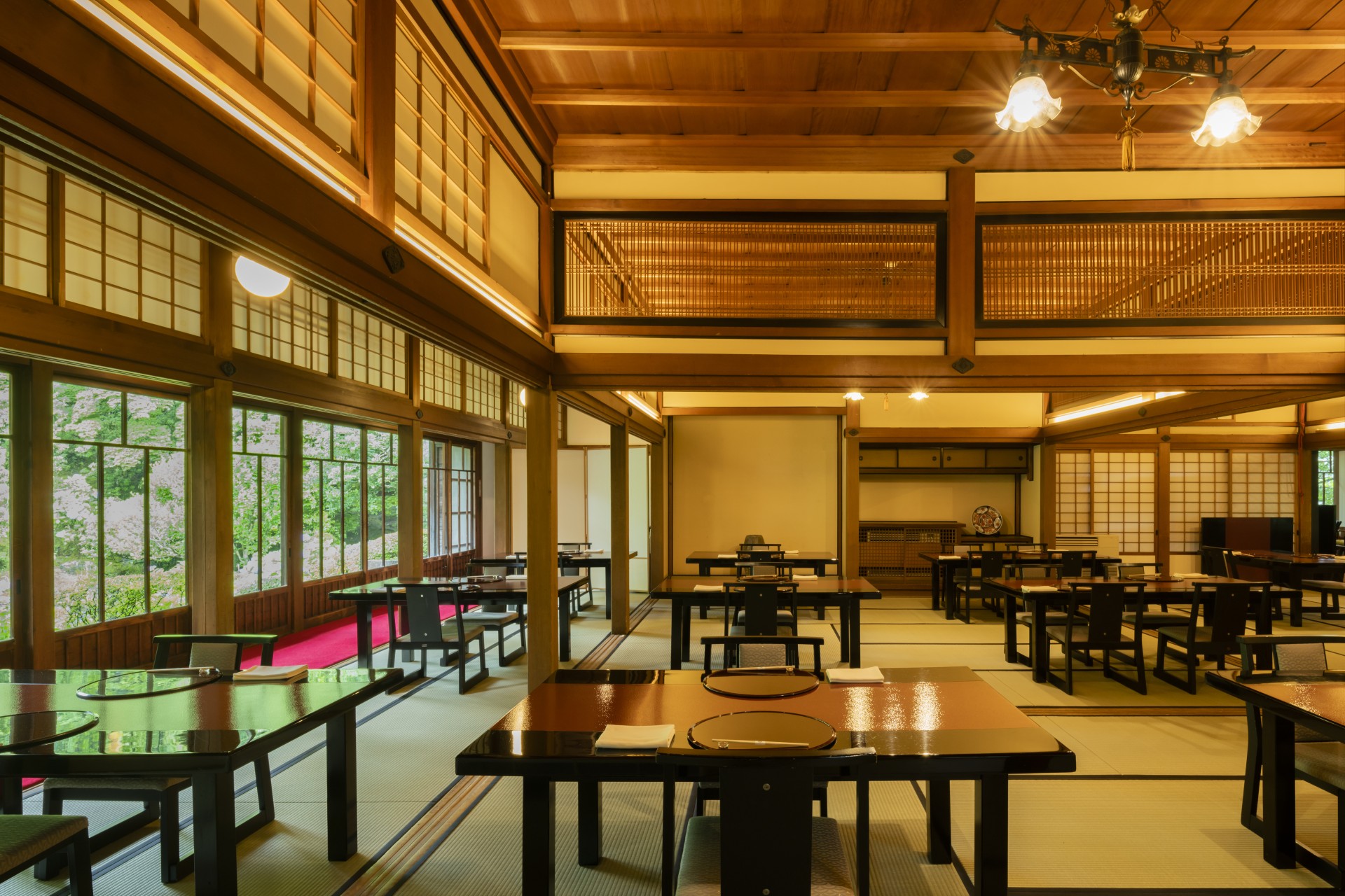 A traditional Japanese dining room with tatami mat flooring, polished low wooden tables with chairs, shoij sliding doors, warm wood paneled ceilings, and large windows offering views of lush greenery outside.
