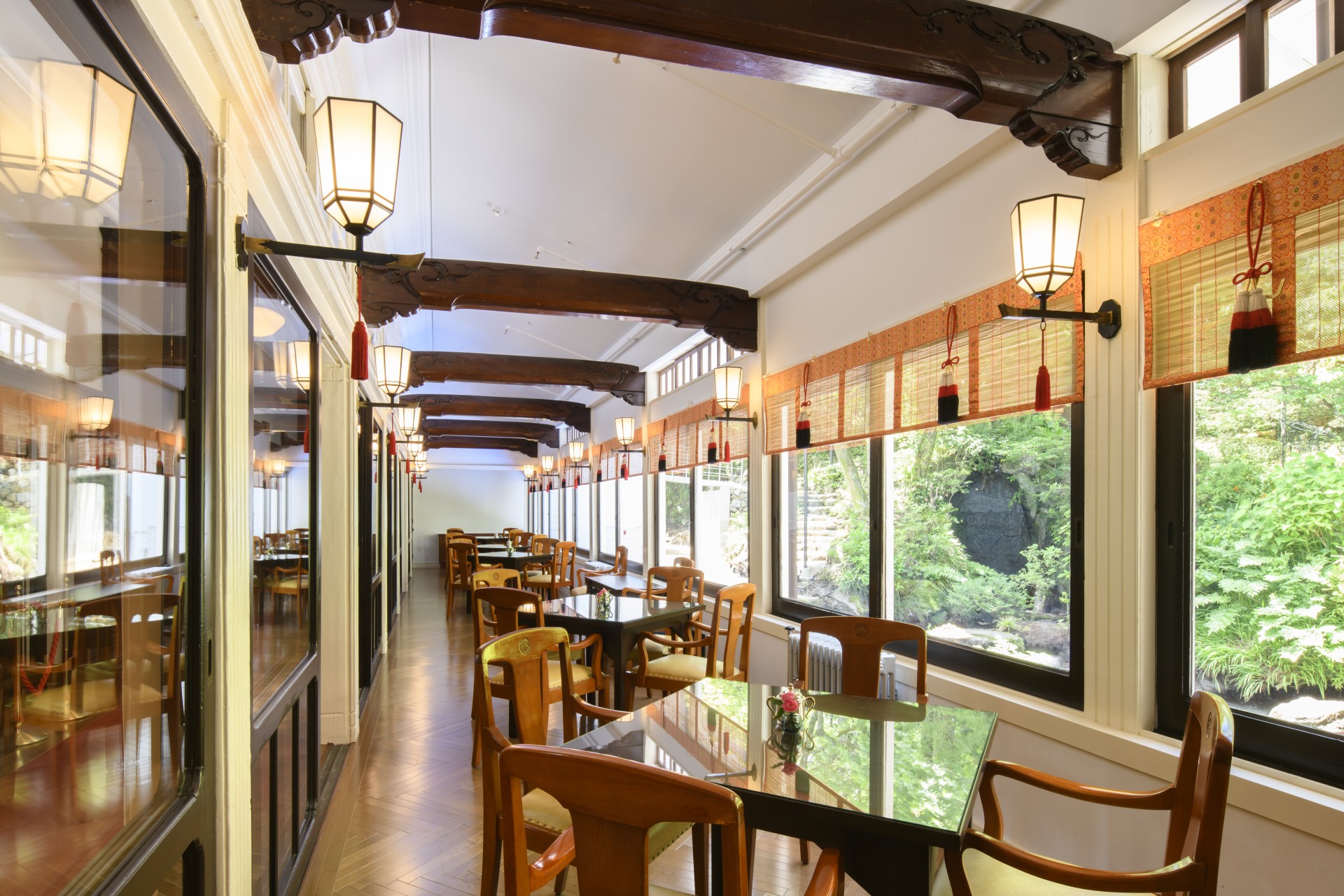 A long, sunlit restaurant hallway featuring small square dining tables with wooden armchairs along a line of large windows. The interior includes dark carved wooden ceiling beams, traditional Japanese lantern wall sconces, bamboo blinds, and views of a ve