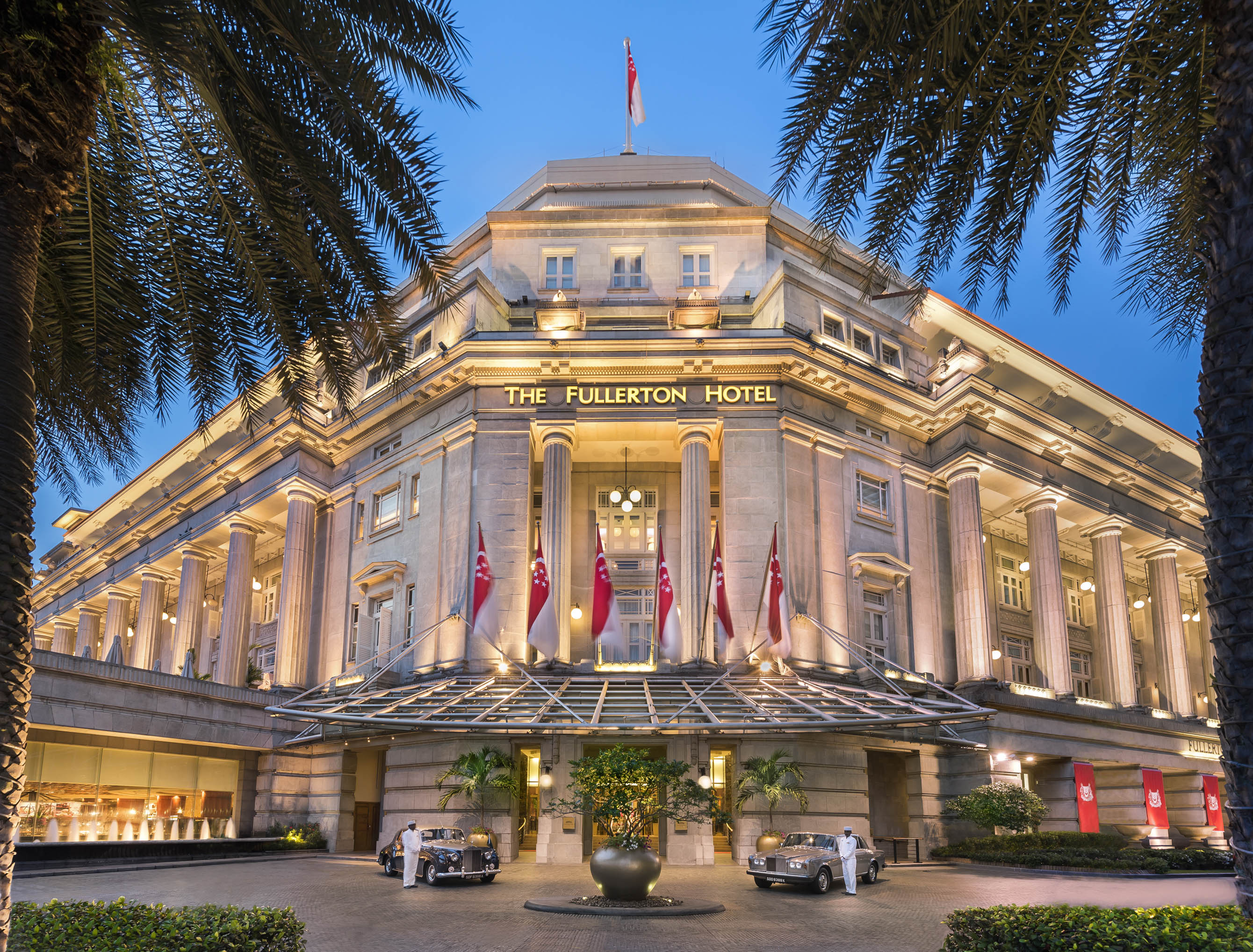 Illuminated grand entrance of The Fullerton Hotel at dusk, featuring classic columns, Singaporean flags, vintage cars parked out front, and palm trees framing the view.