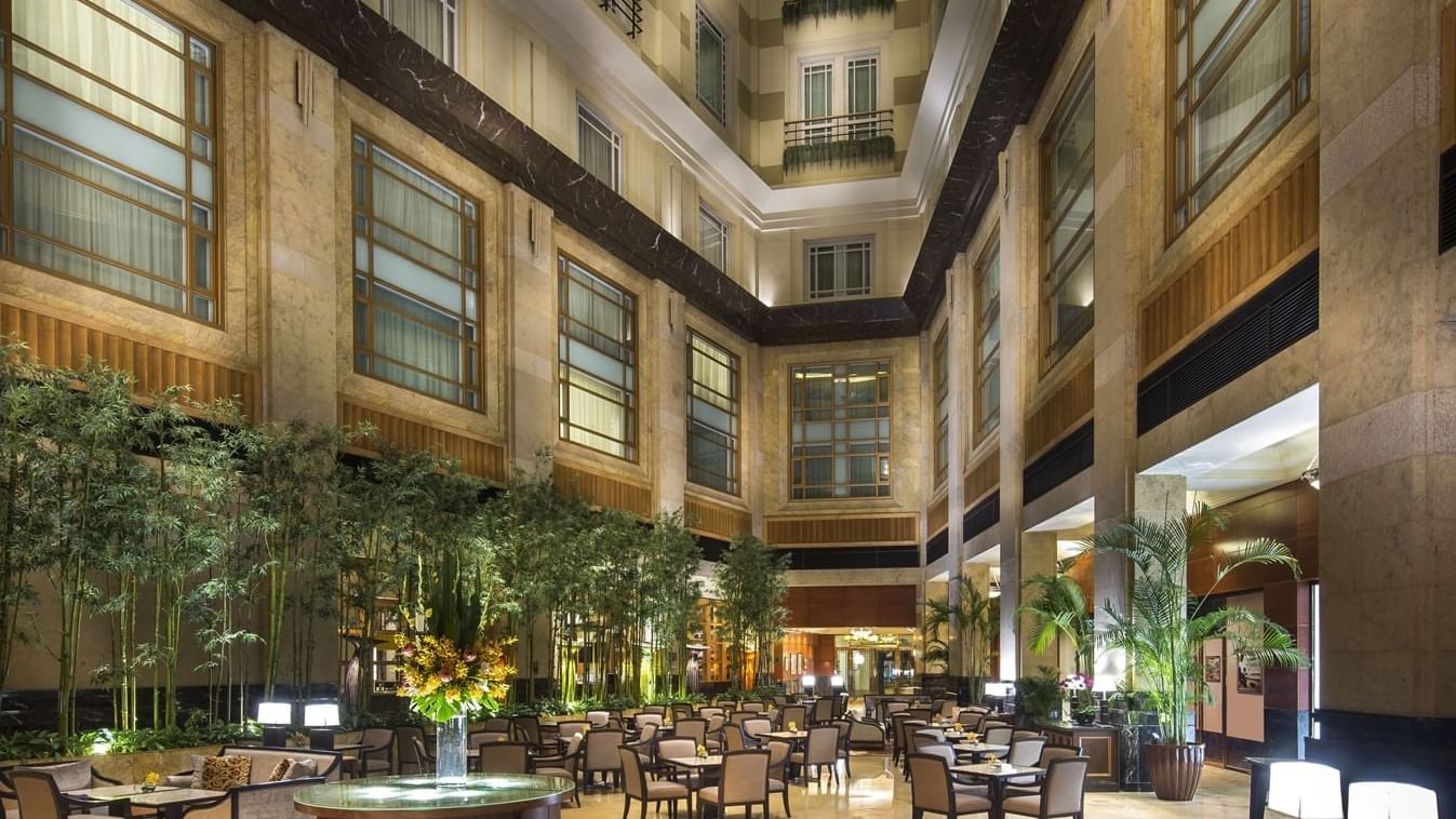 Soaring multi-story indoor atrium restaurant filled with natural light, featuring tall bamboo stalks, lush potted palm trees, marble pillars, and neatly arranged dining tables.