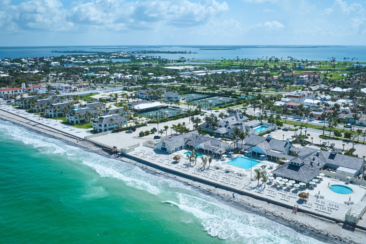 An aerial view of a beachfront resort with white sandy beaches, turquoise ocean waters, swimming pools, lounge chairs, tennis courts, and surrounding coastal buildings.
