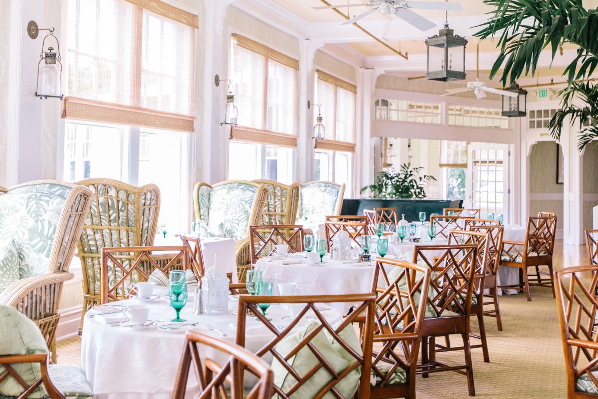 A bright, airy dining room set with white tablecloths, green glassware, bamboo-style wooden chairs, wicker wingback armchairs with palm leaf cushions, large windows with woven shades, and hanging lanterns.