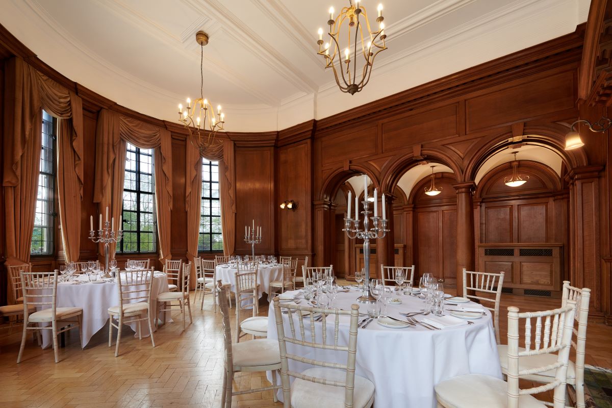 A grand, wood-paneled banquet room featuring round dining tables set with crisp white tablecloths, silver candelabras, and white Chiavari chairs, framed by arched entryways, tall draped windows, and chandeliers hanging from a high white ceiling.