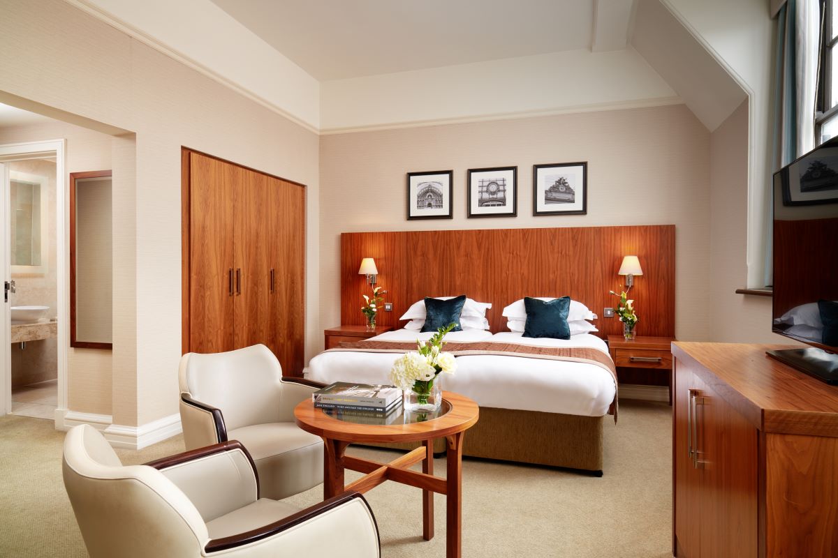 An elegant hotel bedroom featuring a warm wood headboard spanning two twin beds with white linens and dark blue throw pillows, framed black-and-white photos above, two cream armchairs around a round wooden coffee table, and warm wood built-in wardrobes.