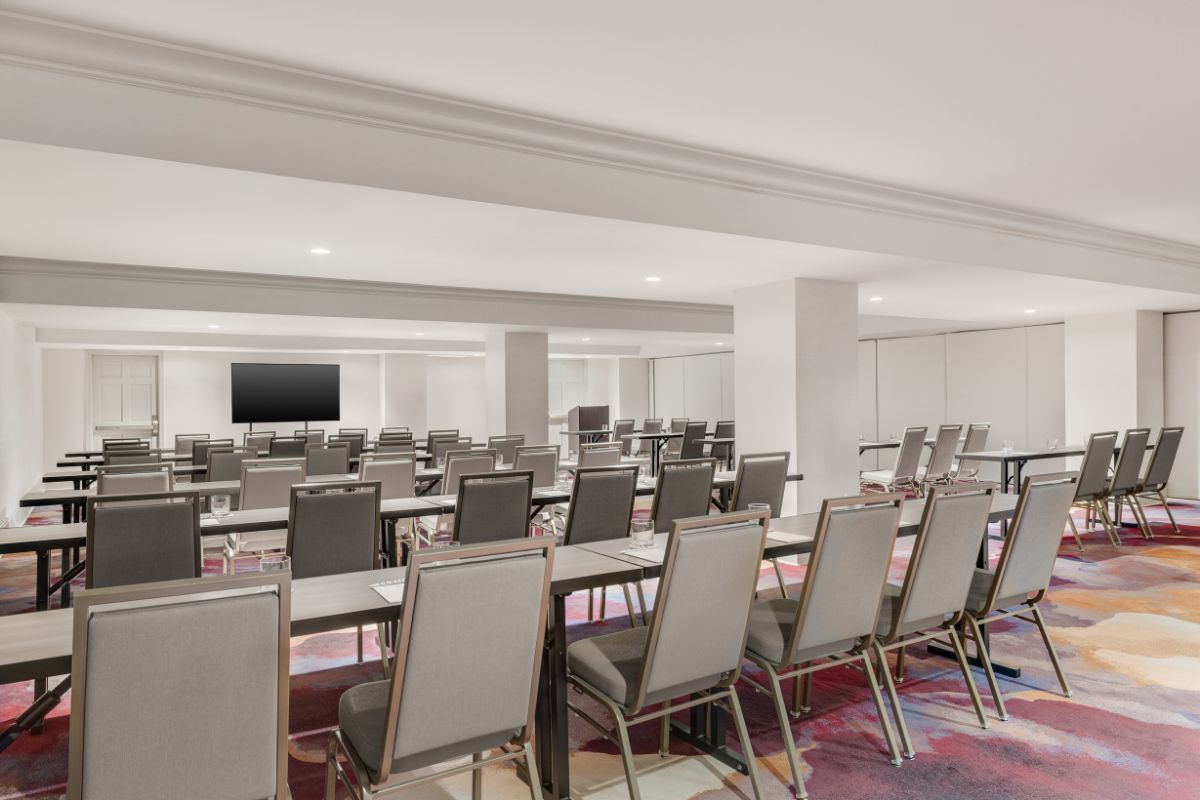 A modern, spacious conference room set up classroom-style with grey padded chairs, dark narrow tables, bright overhead lighting, colorful patterned carpet, and a presentation screen on the wall.