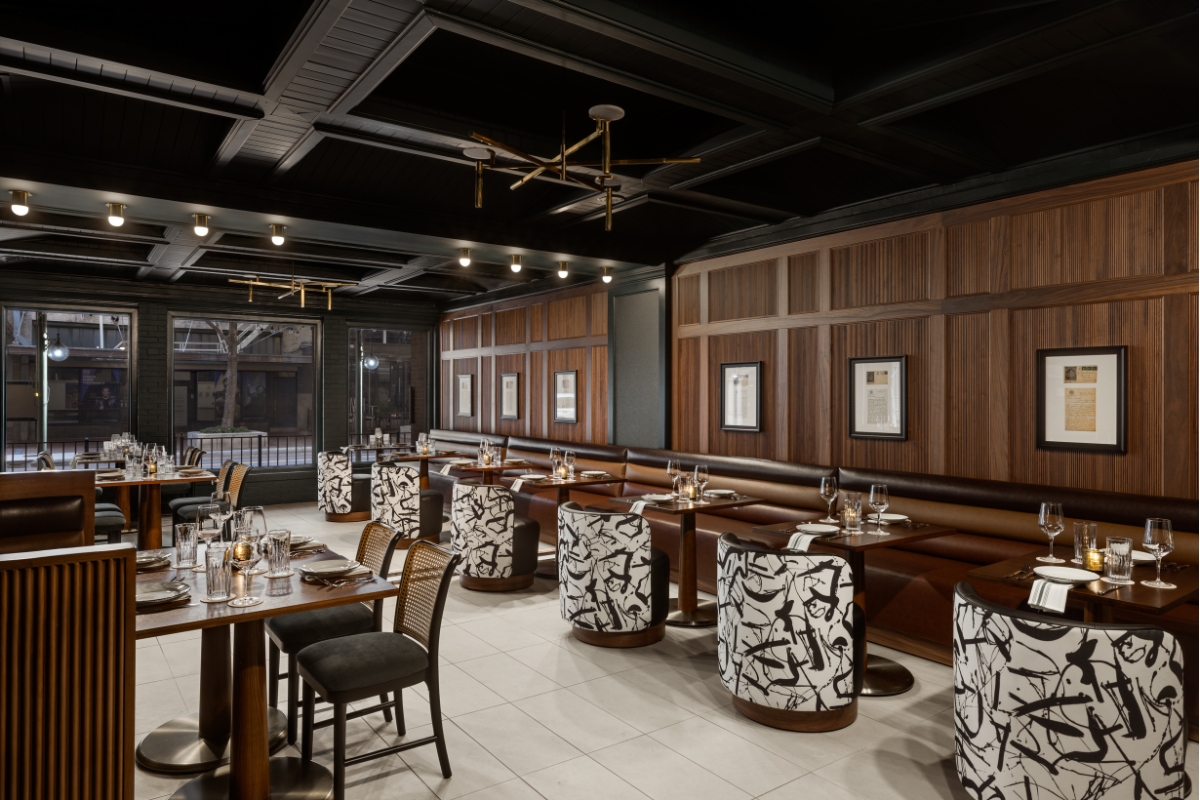 A stylish, dark-ceilinged restaurant interior featuring warm wood-paneled walls, long brown leather banquettes, small wooden dining tables, and bold black-and-white patterned tub chairs.