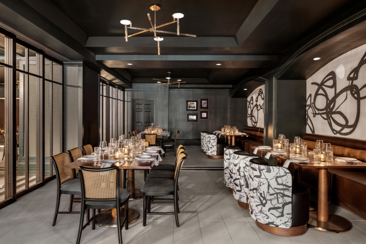 A moody, upscale restaurant space featuring dark ceilings, woven-back dining chairs set along long wooden tables, leather banquettes, abstract wall murals, and modern gold light fixtures.