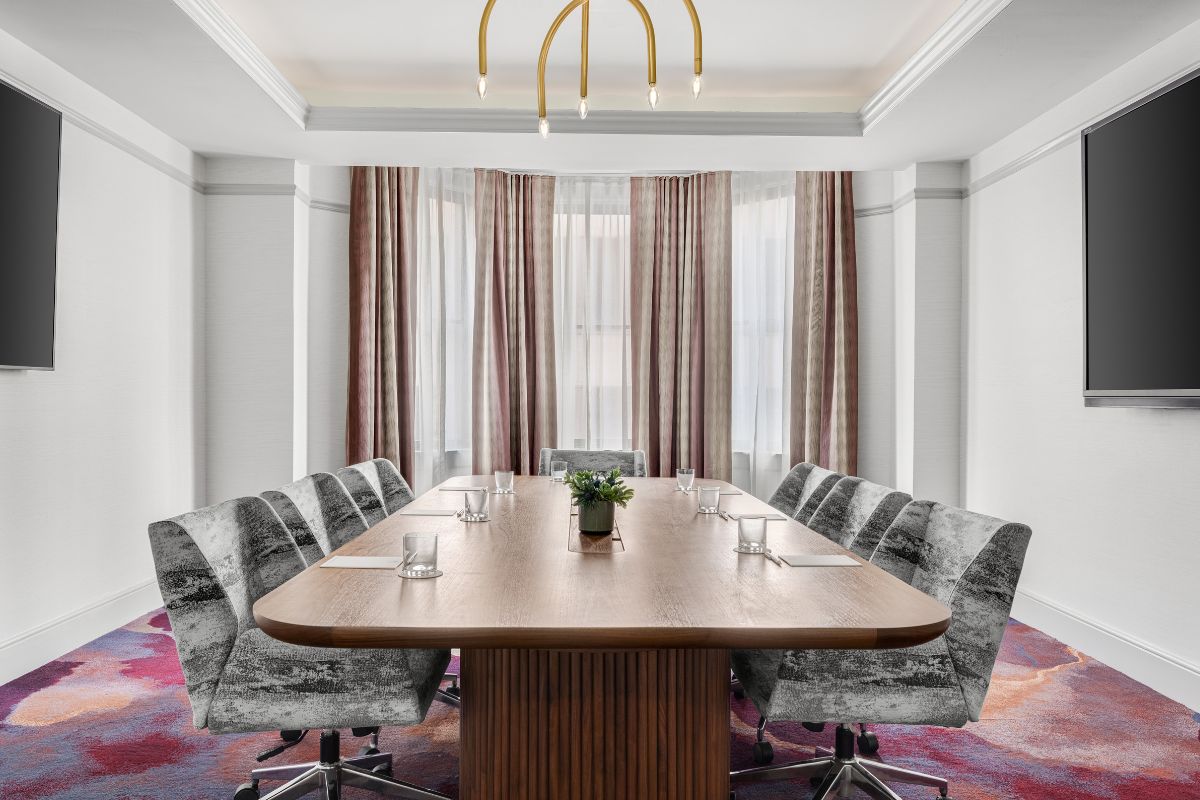 A modern executive boardroom featuring a long wooden conference table, grey textured velvet office chairs, vibrant purple and pink carpeting, a gold arch chandelier, and large bay windows with striped drapes.