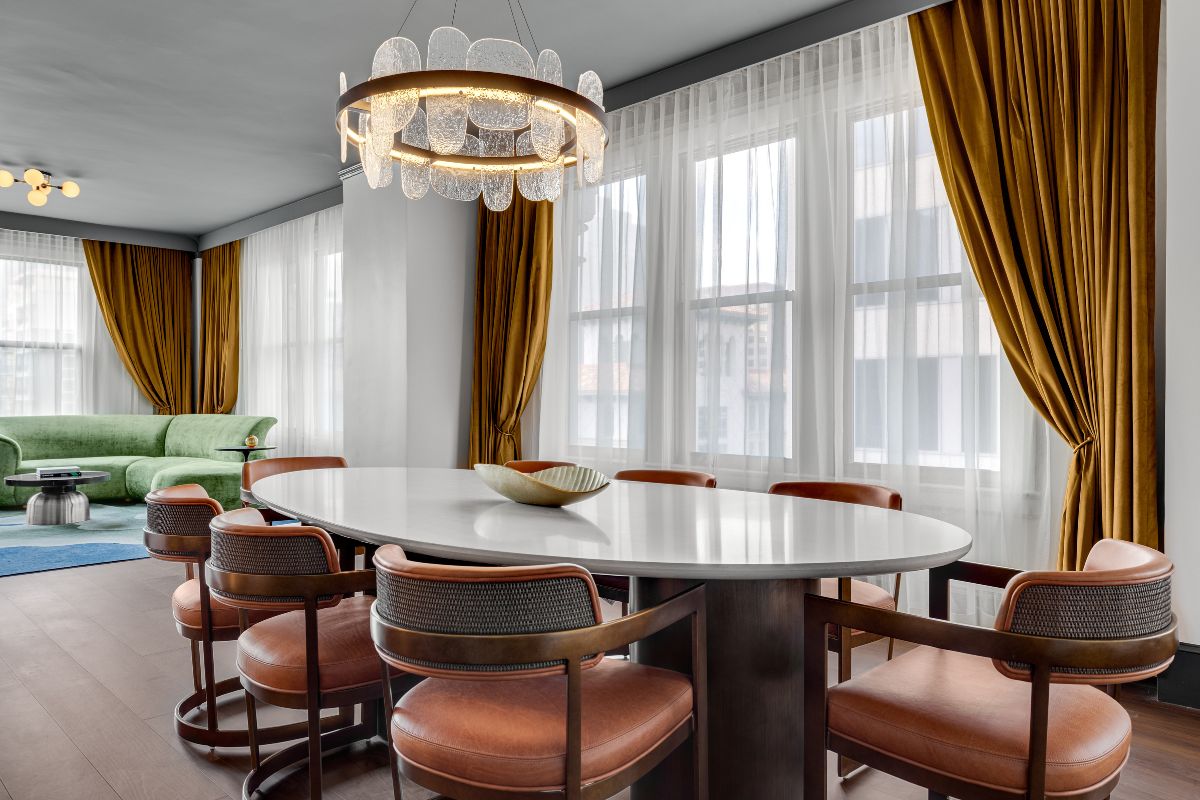 A modern, chic hotel suite dining area featuring a oval white table, brown leather armchairs, a glass ring chandelier, mustard yellow curtains, and a view into an adjacent lounge with a green sofa.