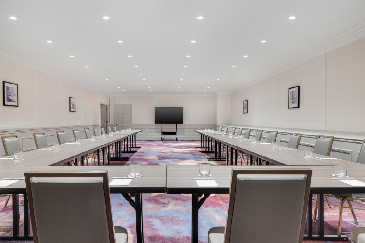 A hotel meeting room arranged in a large U-shape setup with modern grey wood-grain tables, cushioned chairs, recessed ceiling lighting, colorful abstract carpeting, and a mobile presentation monitor at the front.