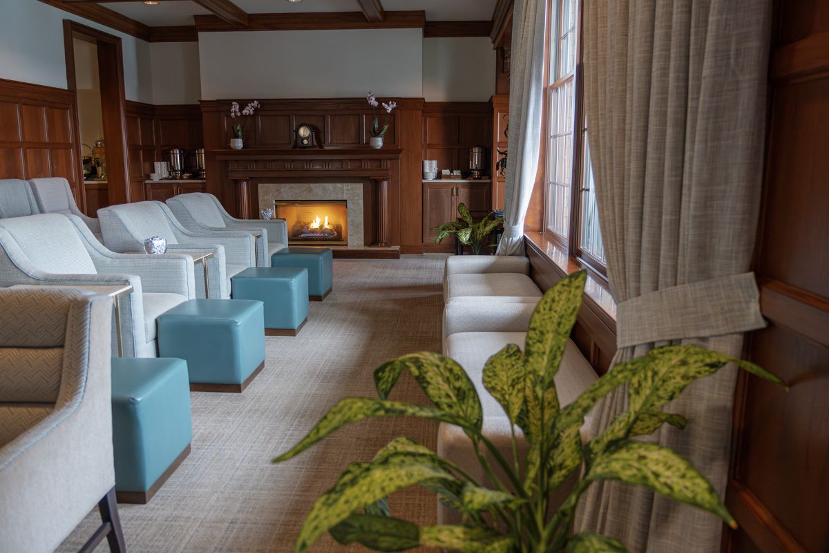 Cozy lounge area with wood-paneled walls, plush armchairs paired with blue ottomans, a glowing fireplace, and large windows with draped curtains.