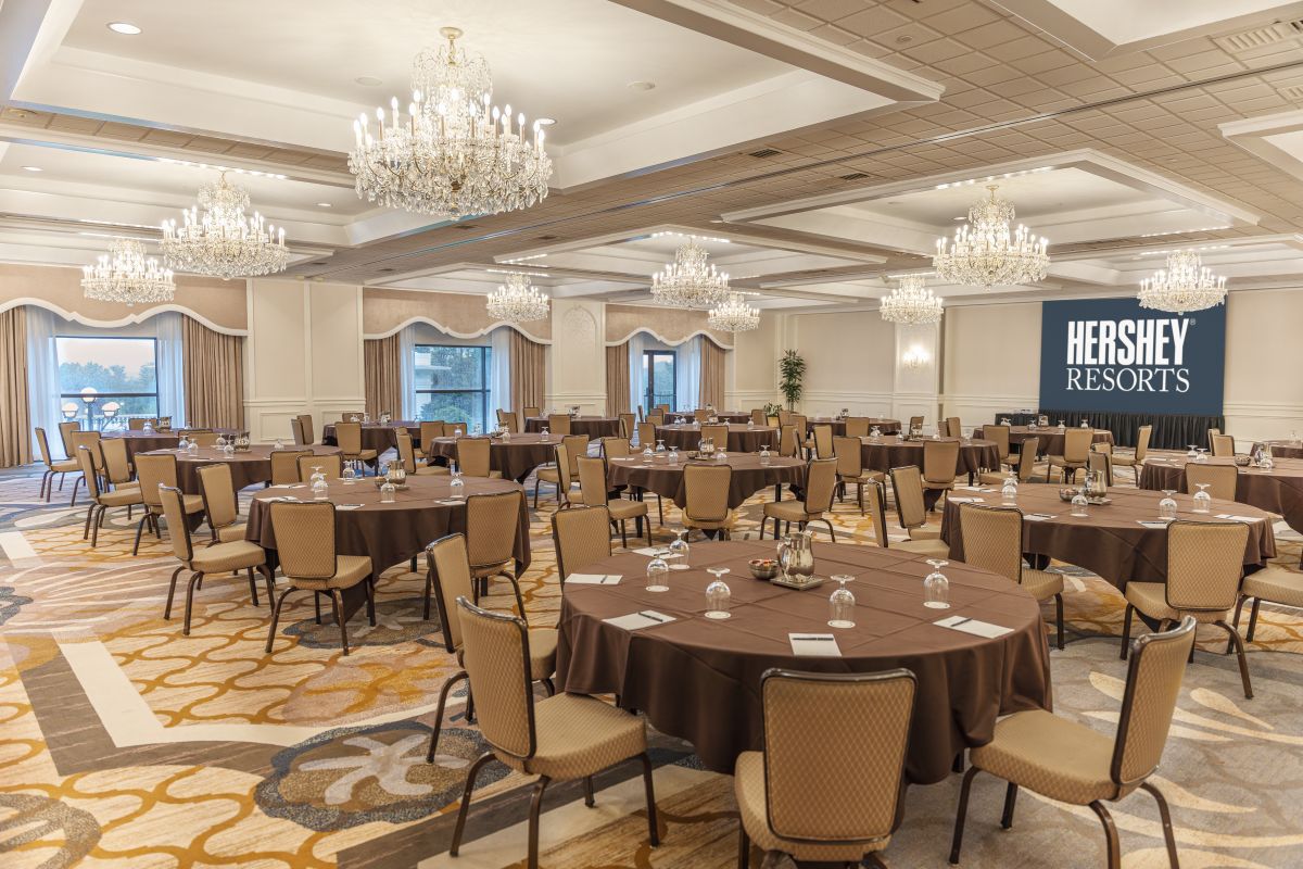 Spacious ballroom configured for a conference, featuring round tables with brown tablecloths, beige chairs, crystal chandeliers, patterned carpeting, and a Hershey Resorts presentation screen.