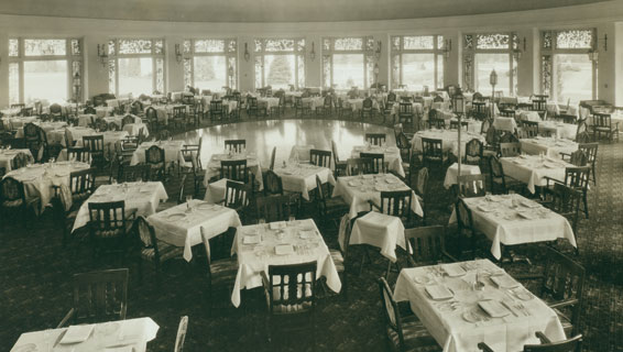 Vintage black-and-white photograph of a large, circular dining room filled with tables set with white tablecloths surrounding a polished wooden dance floor, with large windows line the perimeter.