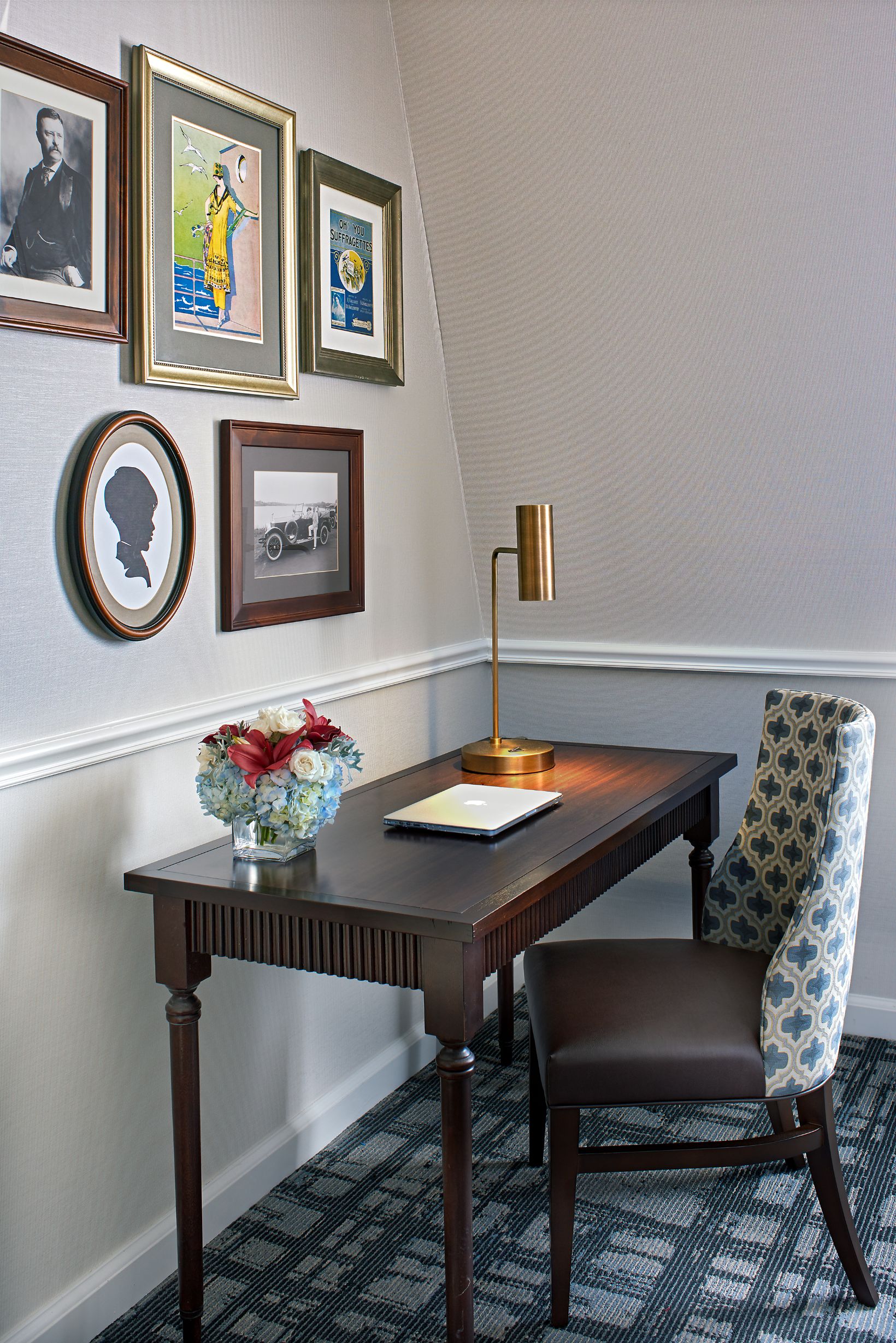 a desk with a lamp and a chair in front of a wall with pictures