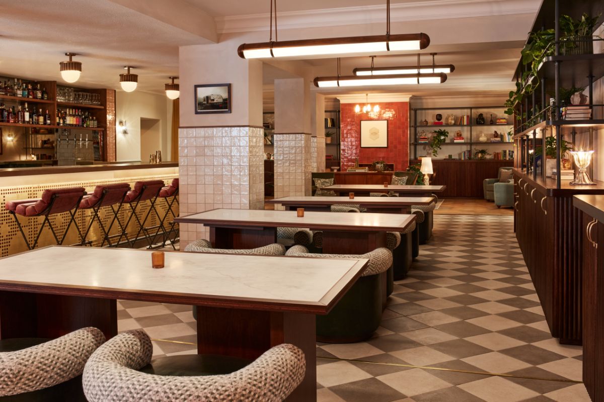 A warm, mid-century modern lounge and bar featuring long white-topped wooden tables paired with textured tweed armchairs, checkered tile flooring, cylindrical pendant lights, glossy white tiled pillars, and a illuminated bar counter lined with red leather