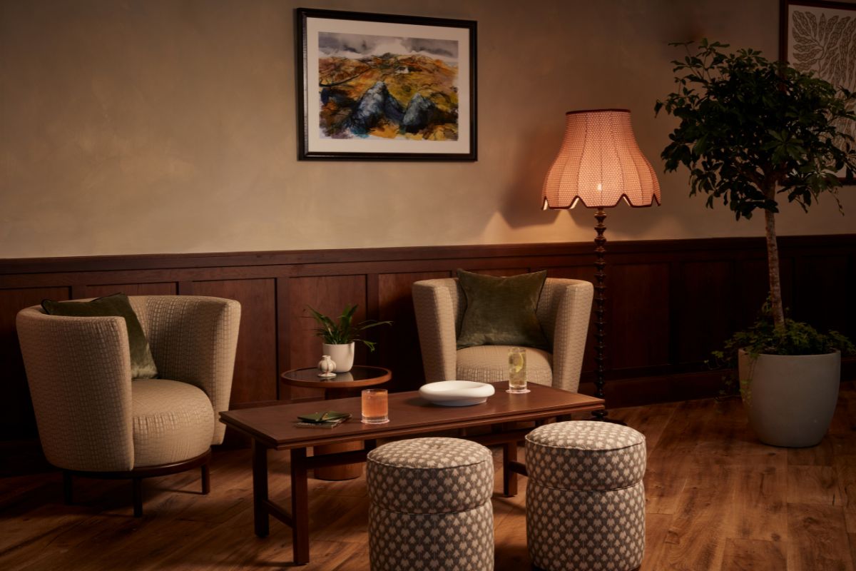 A warm, intimate hotel lounge seating area with two round textured armchairs set against dark wood wall paneling, a low wooden coffee table with matching patterned ottomans, a tall floor lamp with a scalloped shade, and framed artwork.