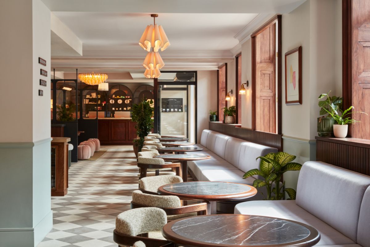 An elegant, modern hotel cafe and seating area featuring a long, light gray upholstered banquette along a window wall, small dark marble round tables, boucle-upholstered tub chairs, layered ceramic pendant lights, and a diagonal checkerboard tile floor le