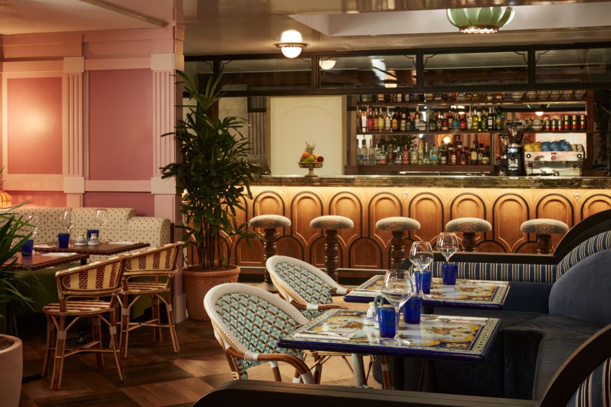 A stylish retro-inspired hotel bar and restaurant area featuring soft pink wall paneling, a wood-front bar counter with arched motifs and cushioned stools, woven bistro-style chairs, and ceramic tiled tables set with blue drinking glasses.