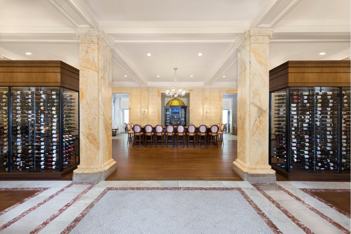 A grand hotel bar entryway framed by large marble columns and two floor-to-ceiling glass wine displays, leading to a long wooden bar lined with high-backed barstools.