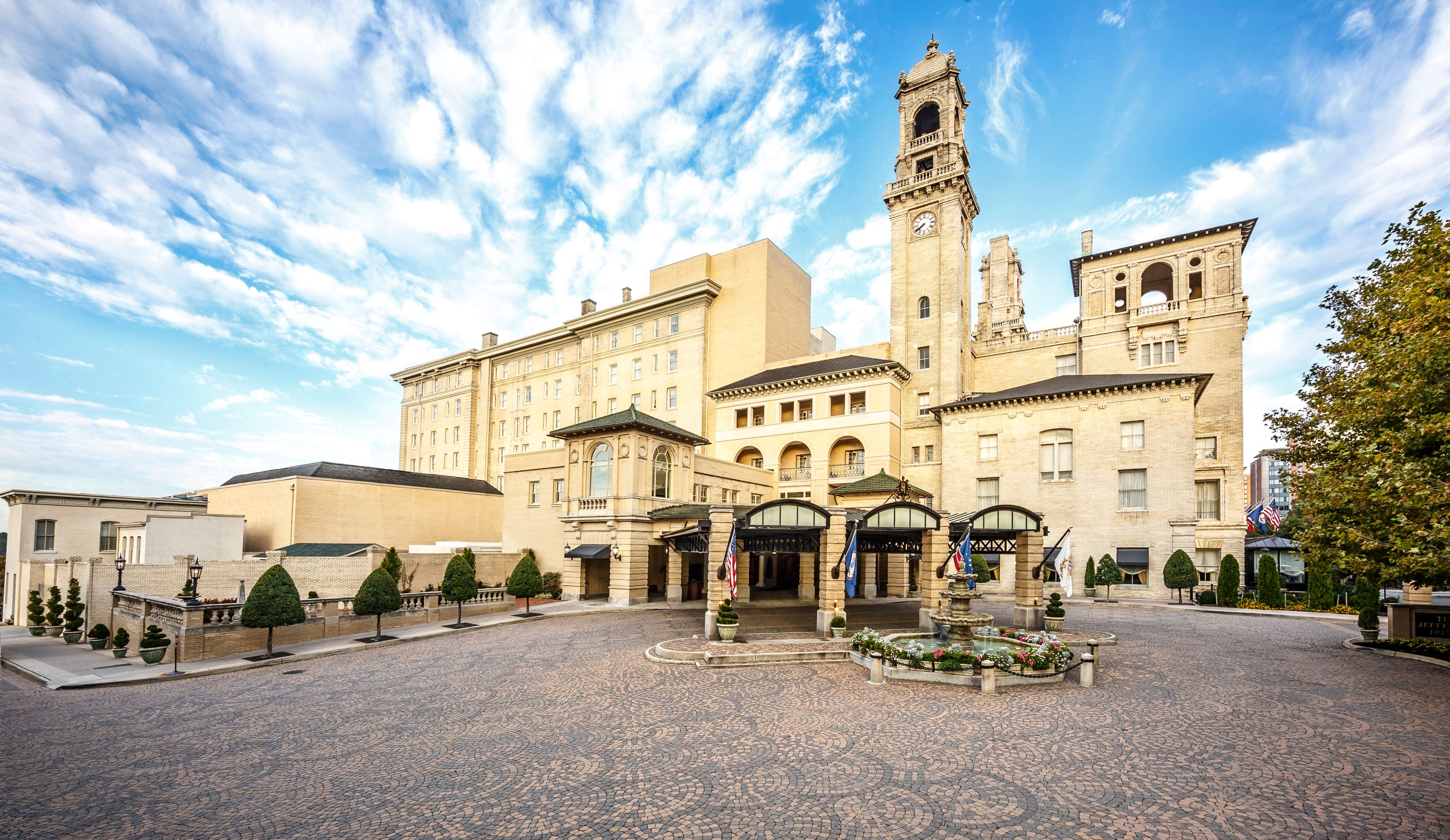 Exterior view of a majestic historic resort hotel with light brick architecture, a prominent clock tower, paved cobblestone driveway, and a central fountain under a bright blue sky.