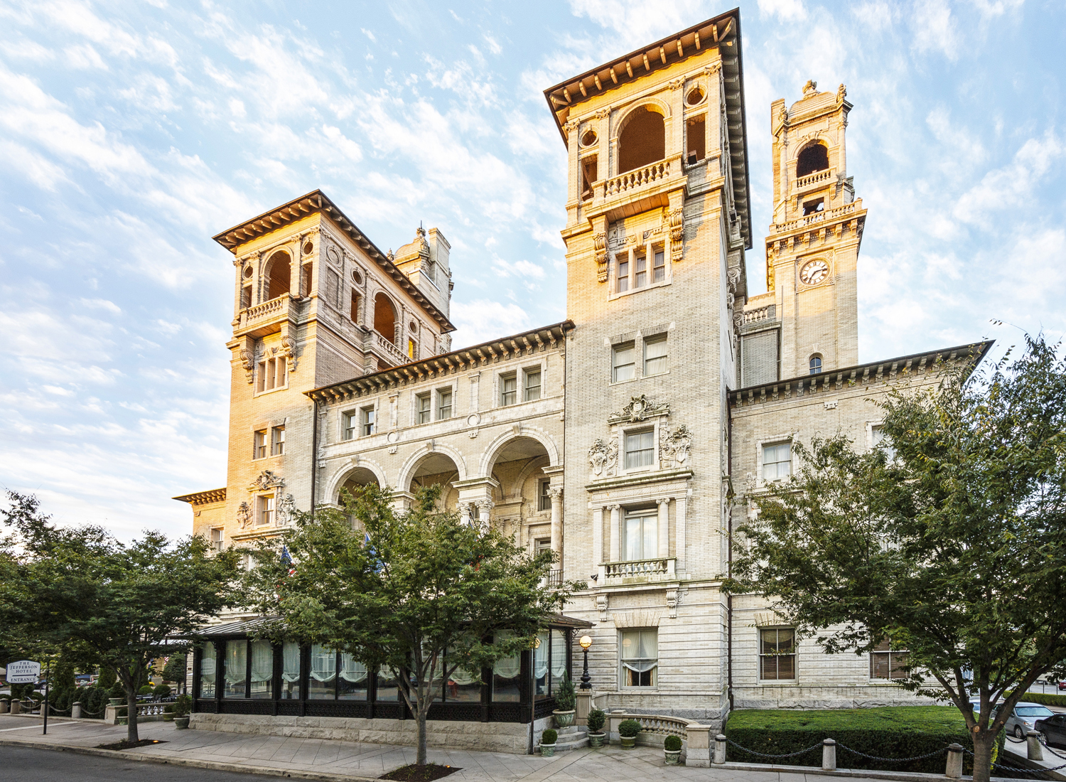 Grand exterior view of a historic hotel with Beaux-Arts architectural towers, stone facades, arched loggias, and lush green trees along the front sidewalk.