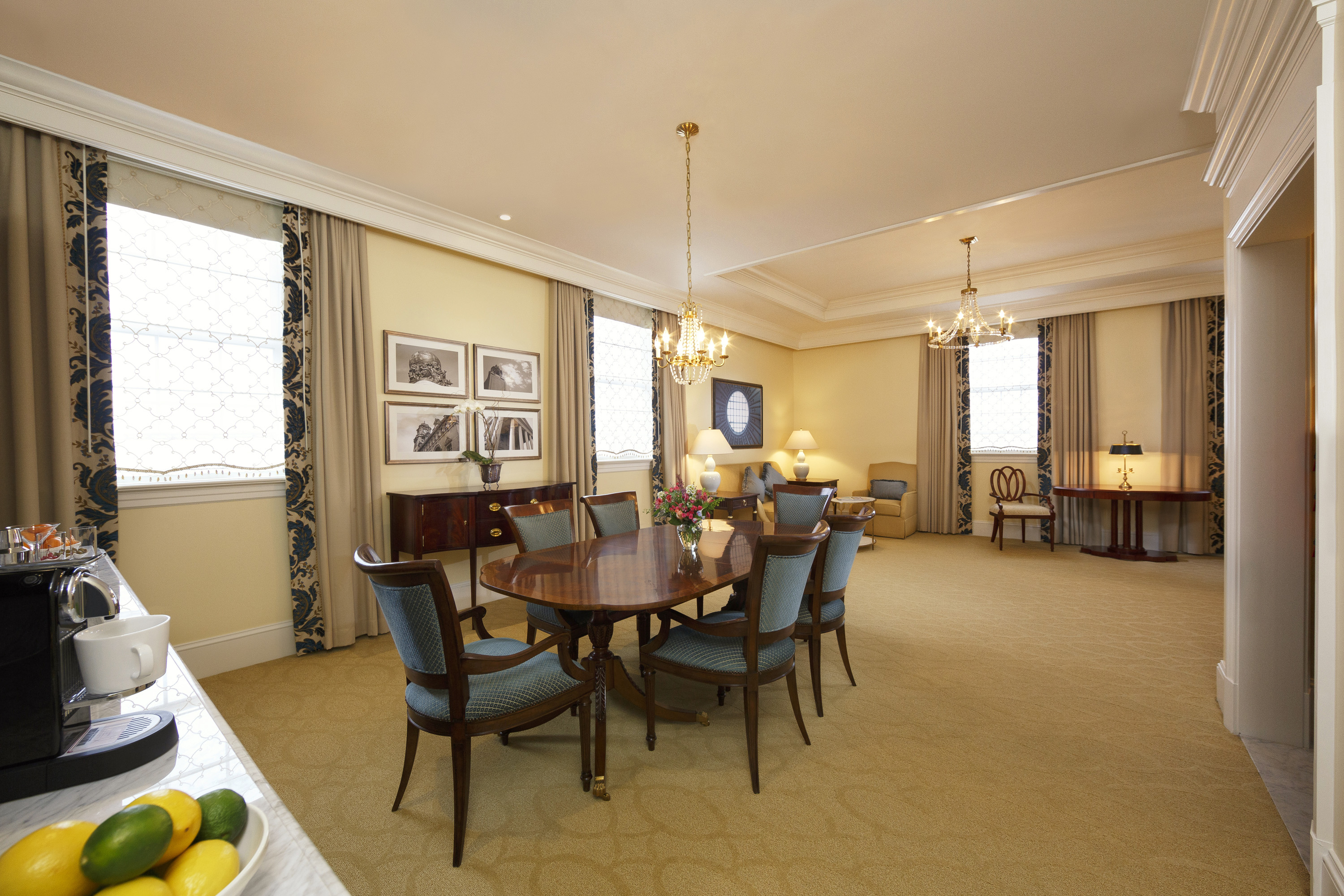 A spacious hotel suite dining area featuring an oval dark wood dining table surrounded by blue upholstered chairs, classic chandeliers, large framed black-and-white photos, and full-length drapes with intricate blue patterns.