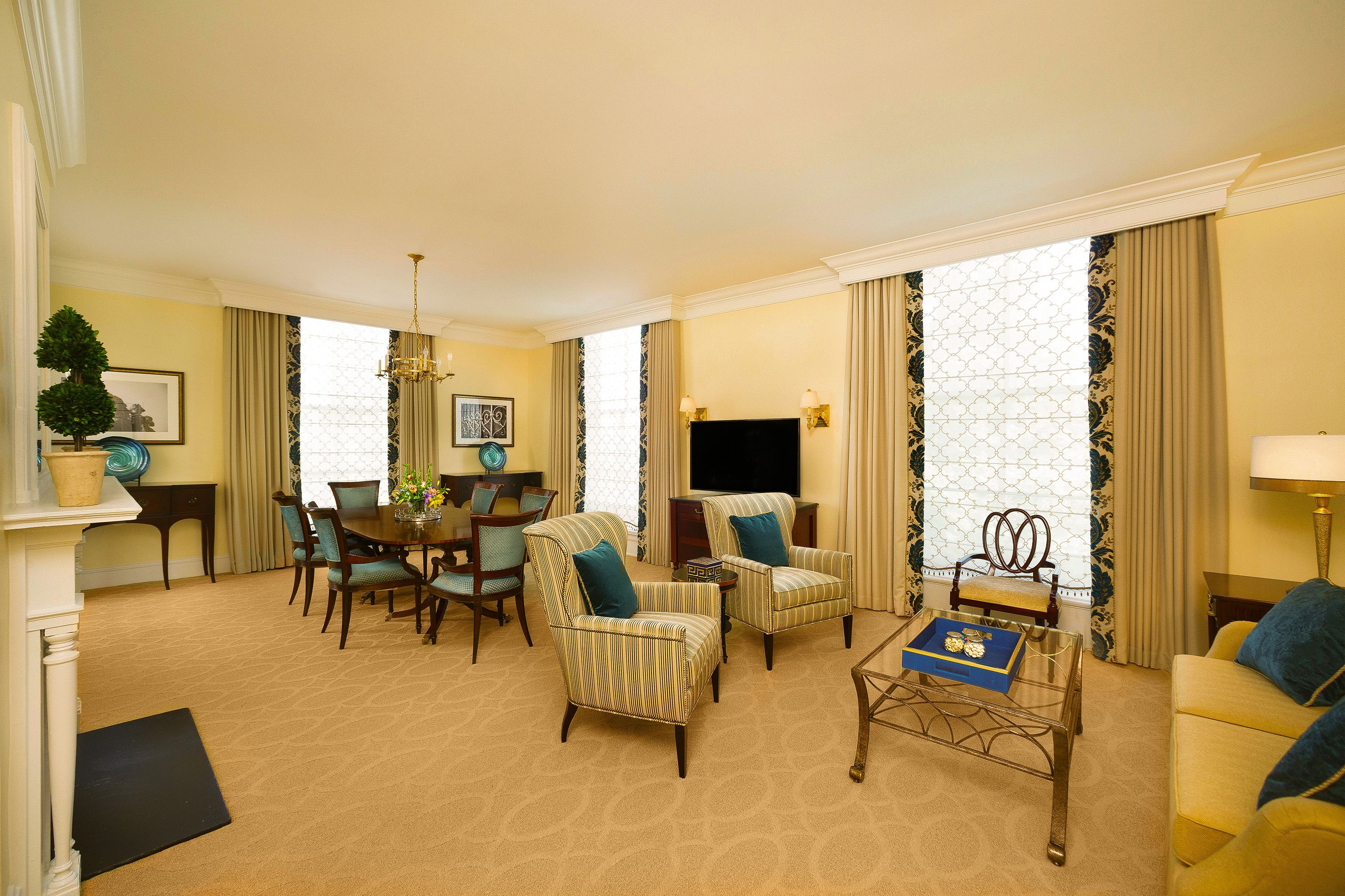 A wide view of an elegant, open-concept hotel suite featuring a living area with striped armchairs, a yellow sofa, and a glass coffee table, alongside a formal dining area with blue-upholstered chairs.
