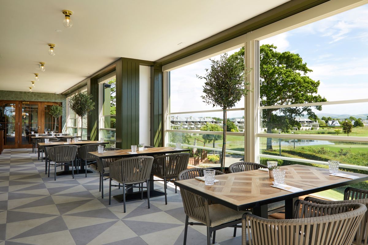 Bright, modern covered dining terrace featuring wooden parquet tables, woven armchair seating, geometric tile flooring, and floor-to-ceiling windows overlooking a green landscape with a lake.