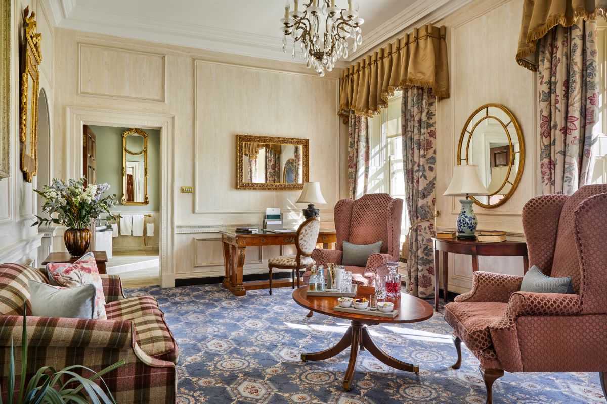 Luxurious traditional suite lounge with blue patterned carpet, plaid sofa, pink wingback armchairs, ornate gold-framed mirrors, wooden writing desk, crystal chandelier, and view into an adjacent bathroom.