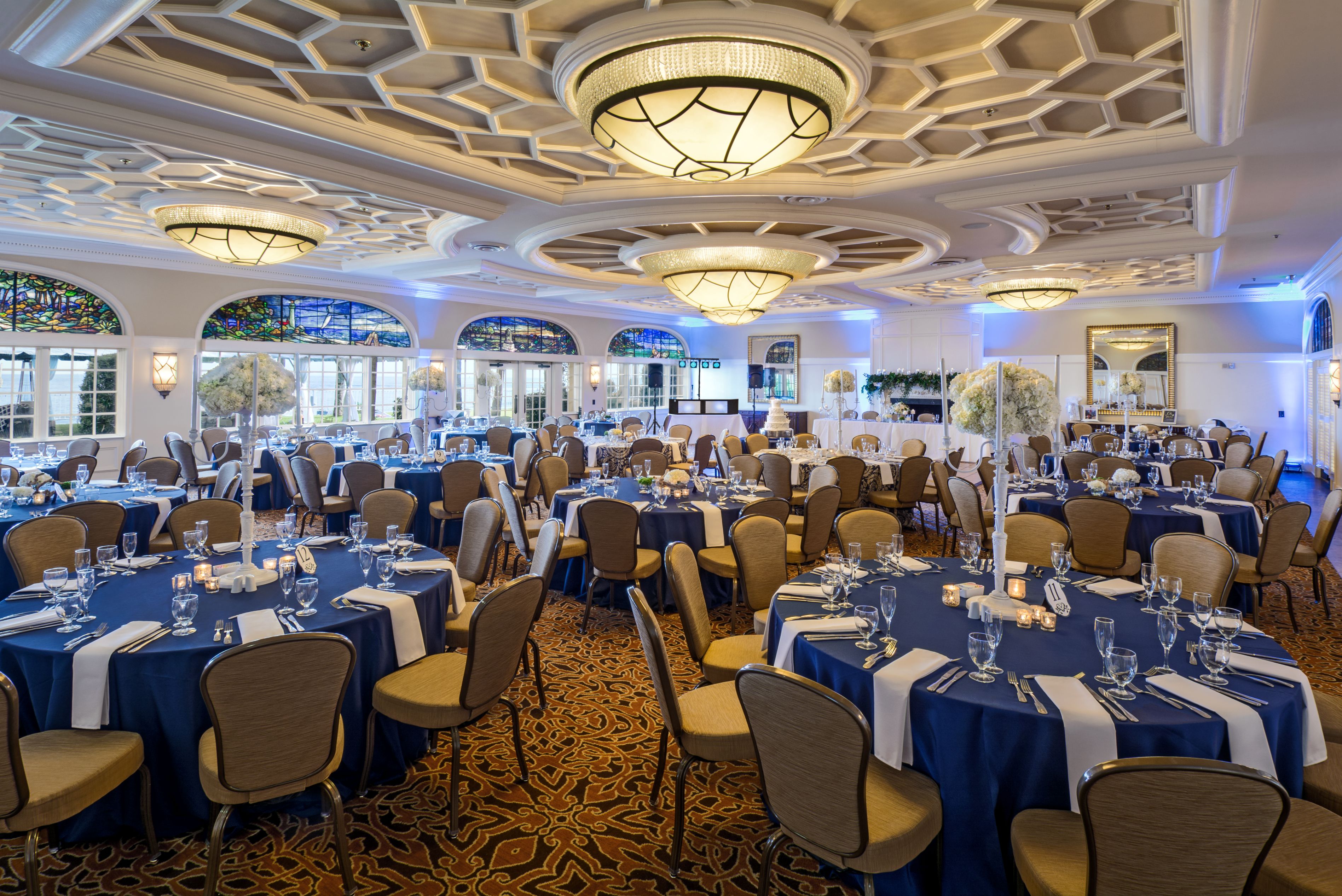Grand event ballroom set up with round tables dressed in deep blue tablecloths, tall white floral centerpieces, beige upholstered chairs, intricate coffered ceiling with circular light fixtures, and stained glass windows.