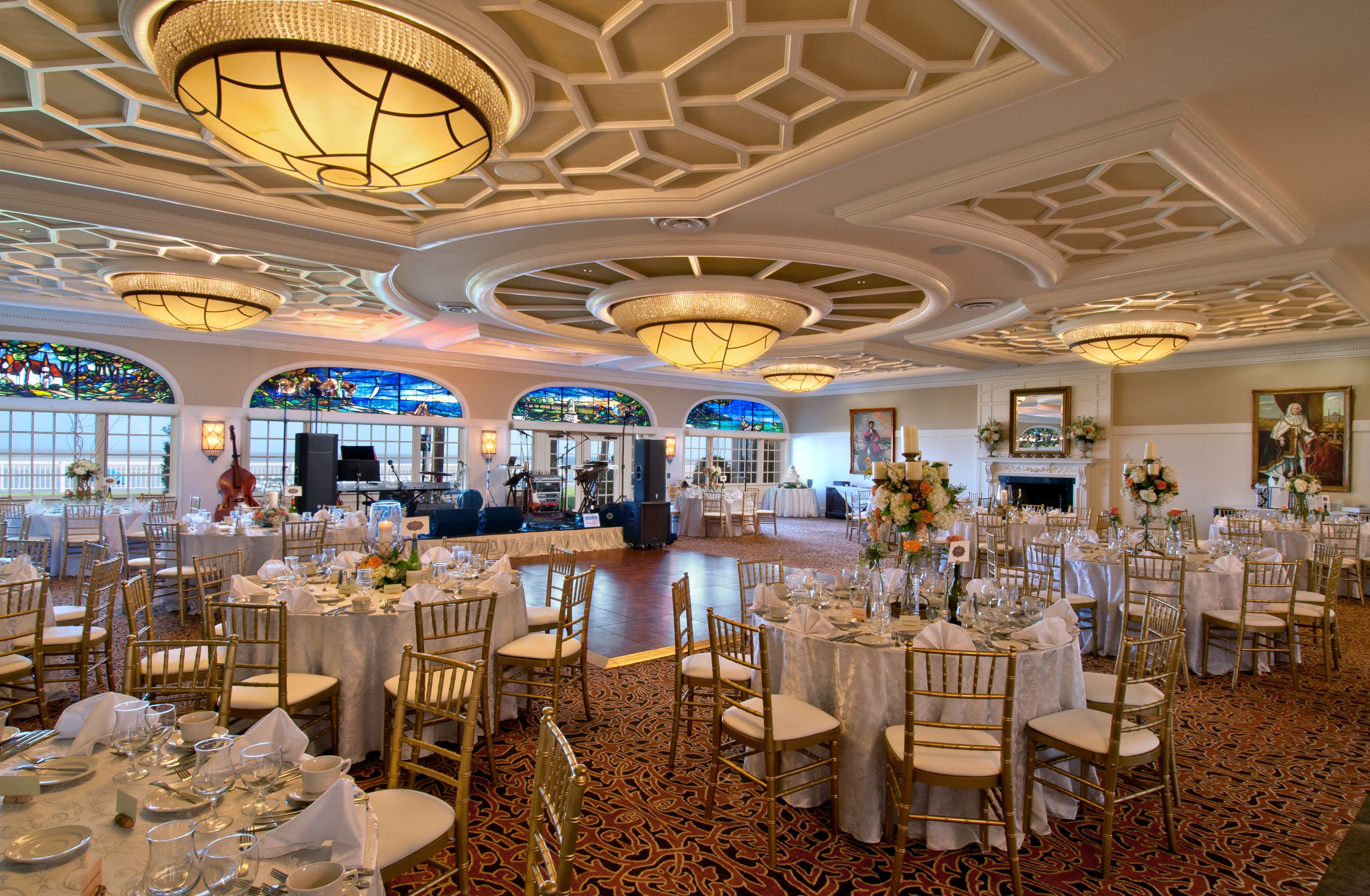 Elegant event ballroom set up for a reception with white round tablecloths, gold Chiavari chairs, floral centerpieces, a dance floor in front of a stage setup, intricate geometric ceiling with flush-mount chandeliers, and stained glass window accents.