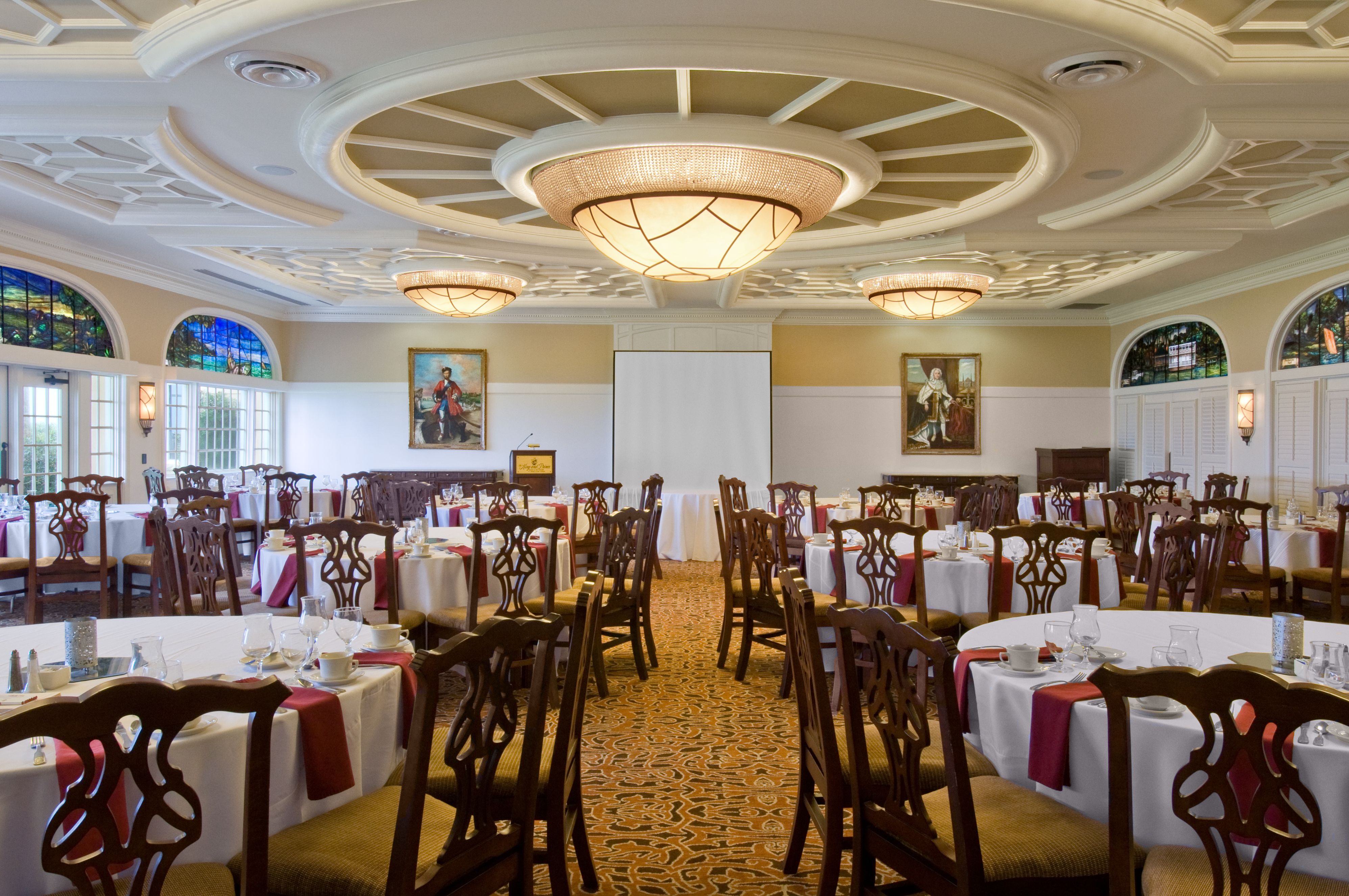 Large banquet room configured for a conference with round tables with white linens and red napkins, carved wooden chairs, a presentation screen and podium at the front, framed portraits, and decorative ceiling chandeliers.