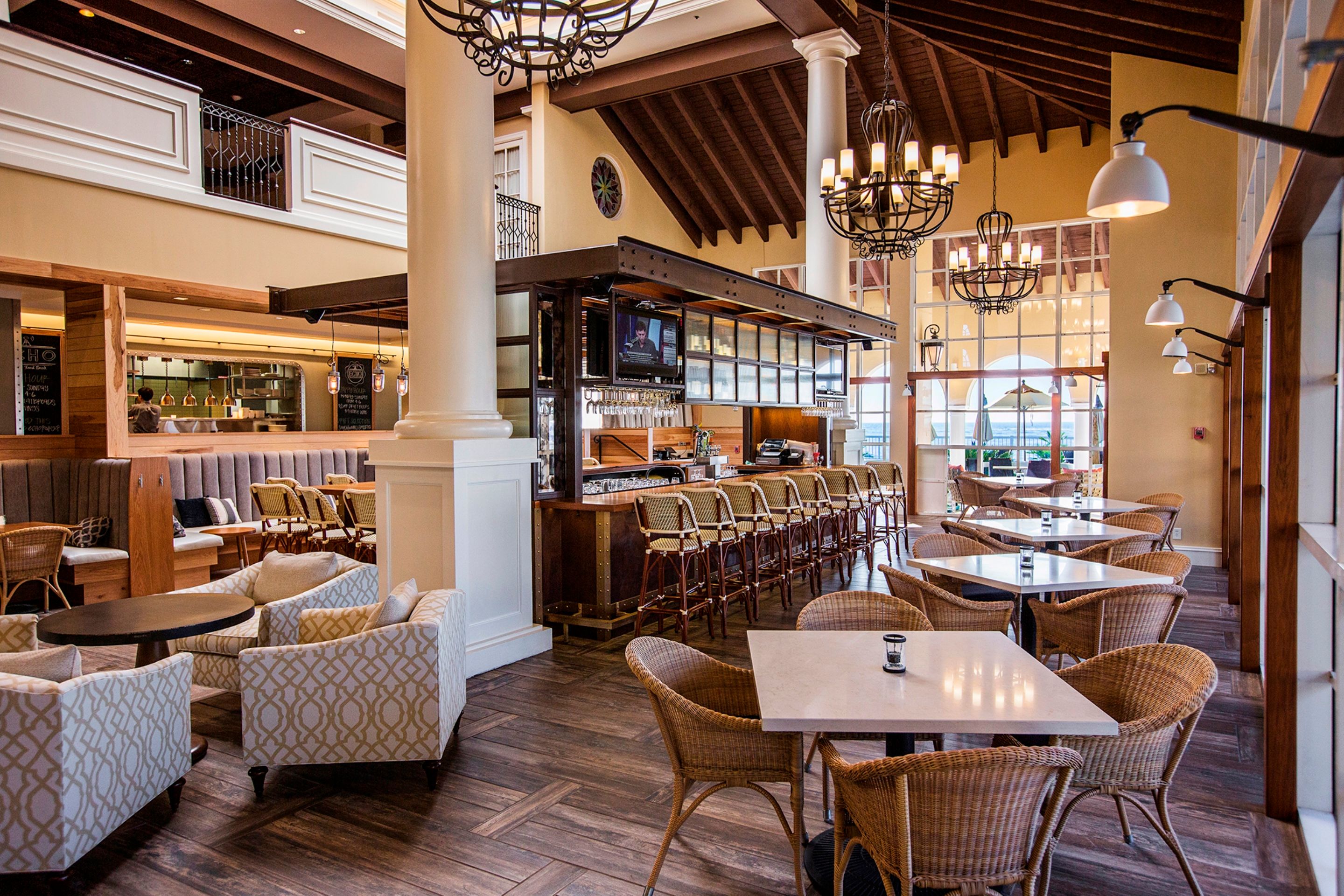 Airy, high-ceilinged hotel restaurant and bar featuring wood-paneled cathedral ceilings, wrought-iron chandeliers, a central island bar with high wicker stools, surrounding dining tables, and plush patterned lounge armchairs.