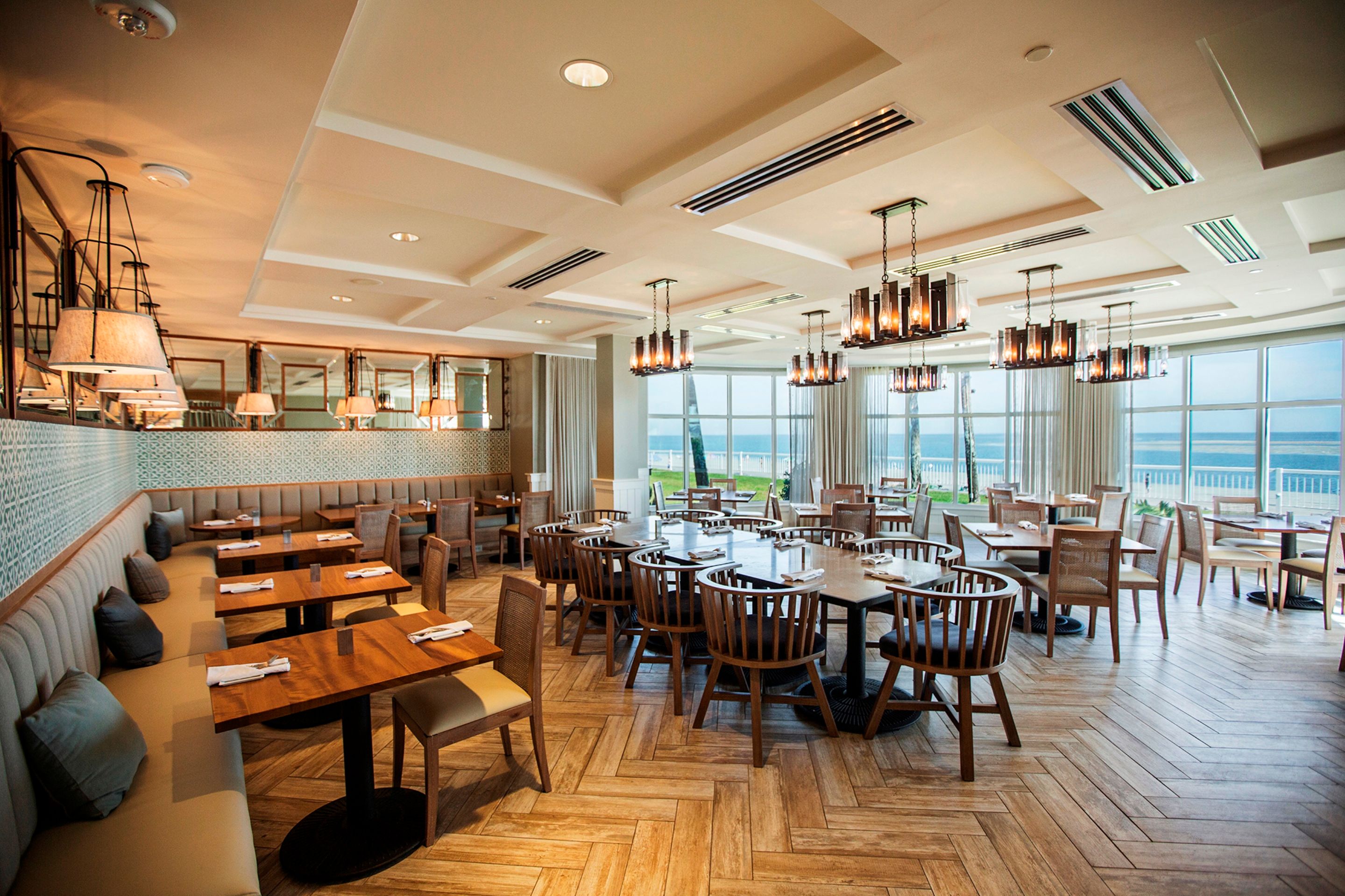 Modern, light-filled oceanfront restaurant with herringbone wood floors, a long banquette sofa lined with pillows, wooden dining tables, woven armchairs, and floor-to-ceiling windows offering views of the ocean.