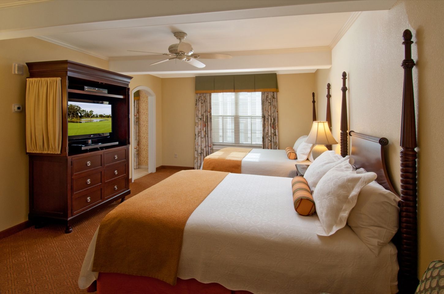 Hotel room featuring two double beds with white linens, gold throw blankets, four-poster wooden bed frames, a wooden armoire with a television, and a window with patterned drapes.