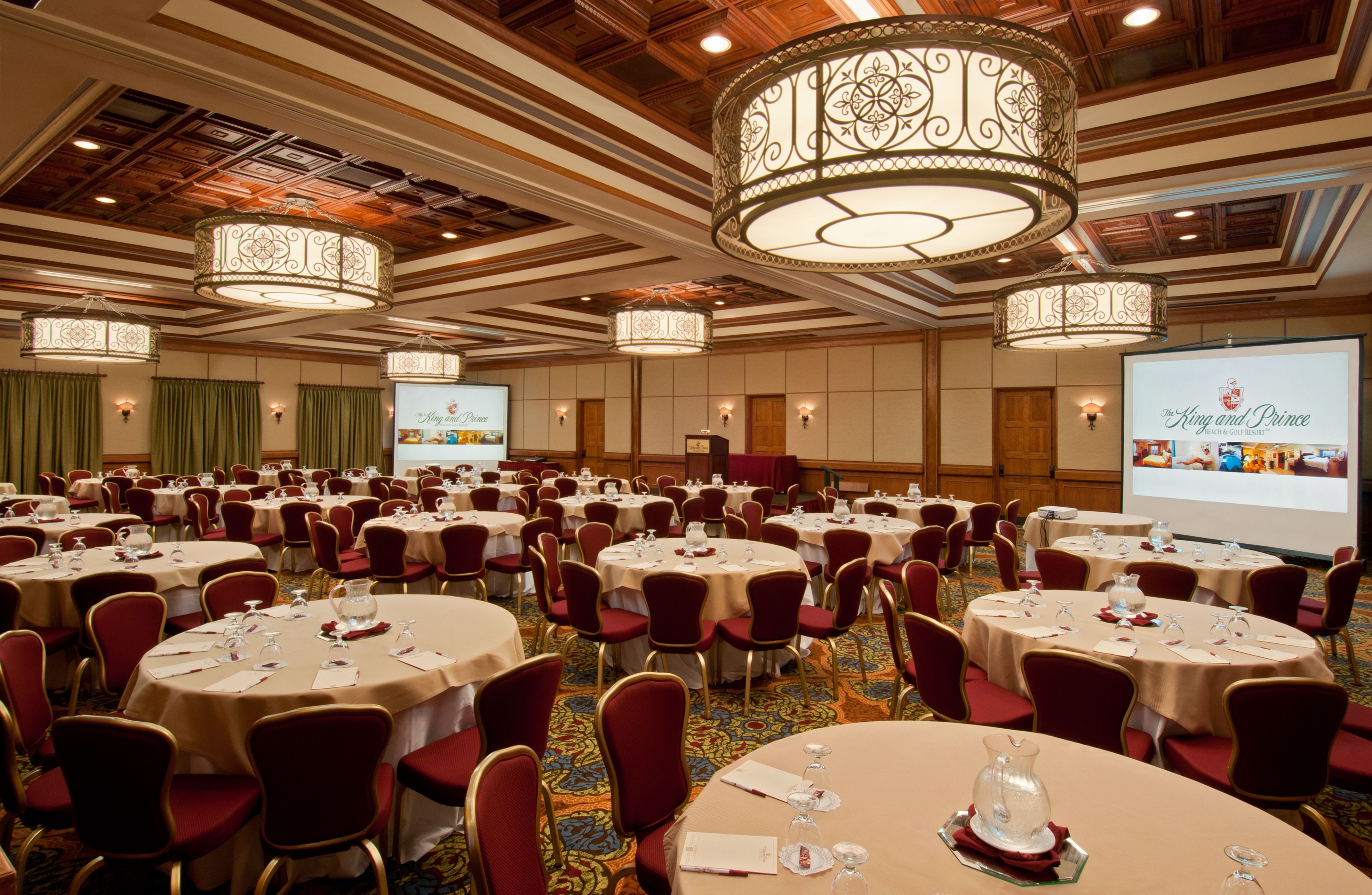 Large conference ballroom set up with round banquet tables topped with beige linens, water pitchers, and place settings, surrounded by maroon padded chairs, featuring large ornate drum chandeliers, coffered ceilings, and two projection screens displaying 