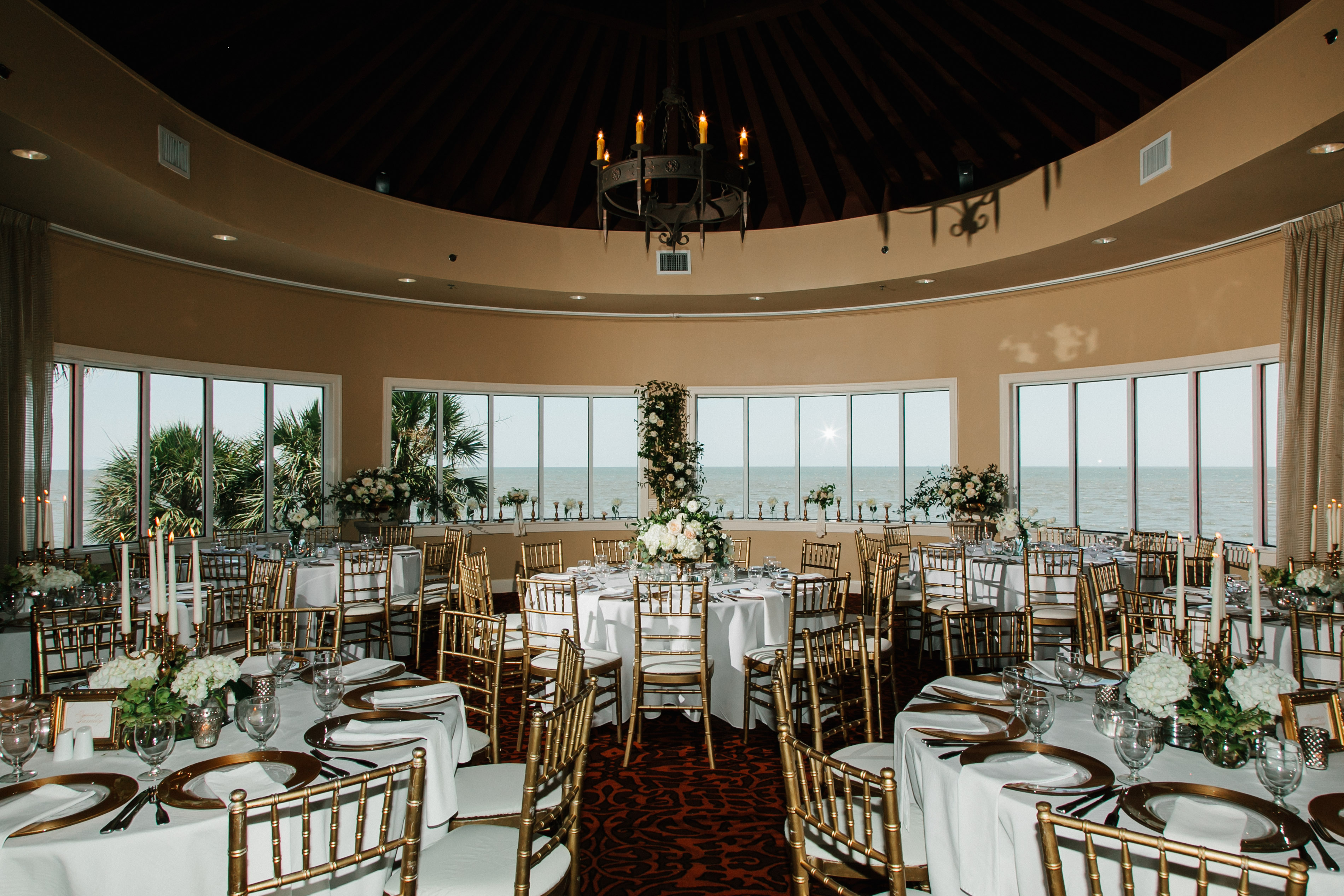 Circular event dining room set up for a wedding reception with round tables, gold Chiavari chairs, elegant floral arrangements, candelabras, panoramic windows overlooking the ocean, and a dark wood vaulted cone ceiling with a chandelier.