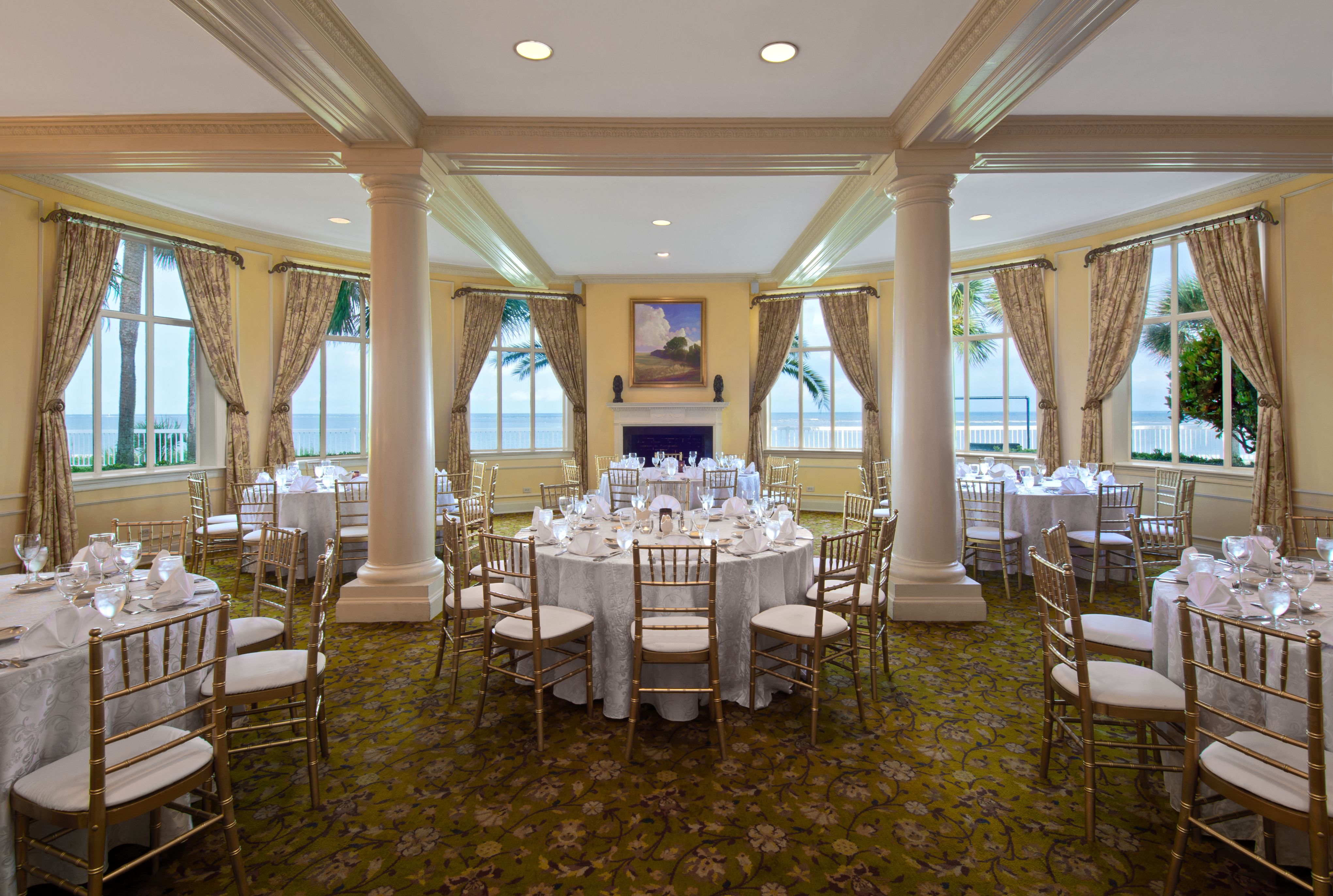 Bright, round event room set for a reception with white damask round tables, gold Chiavari chairs, prominent white support columns, surrounding windows with ocean views and draped curtains, and a central fireplace in the background.