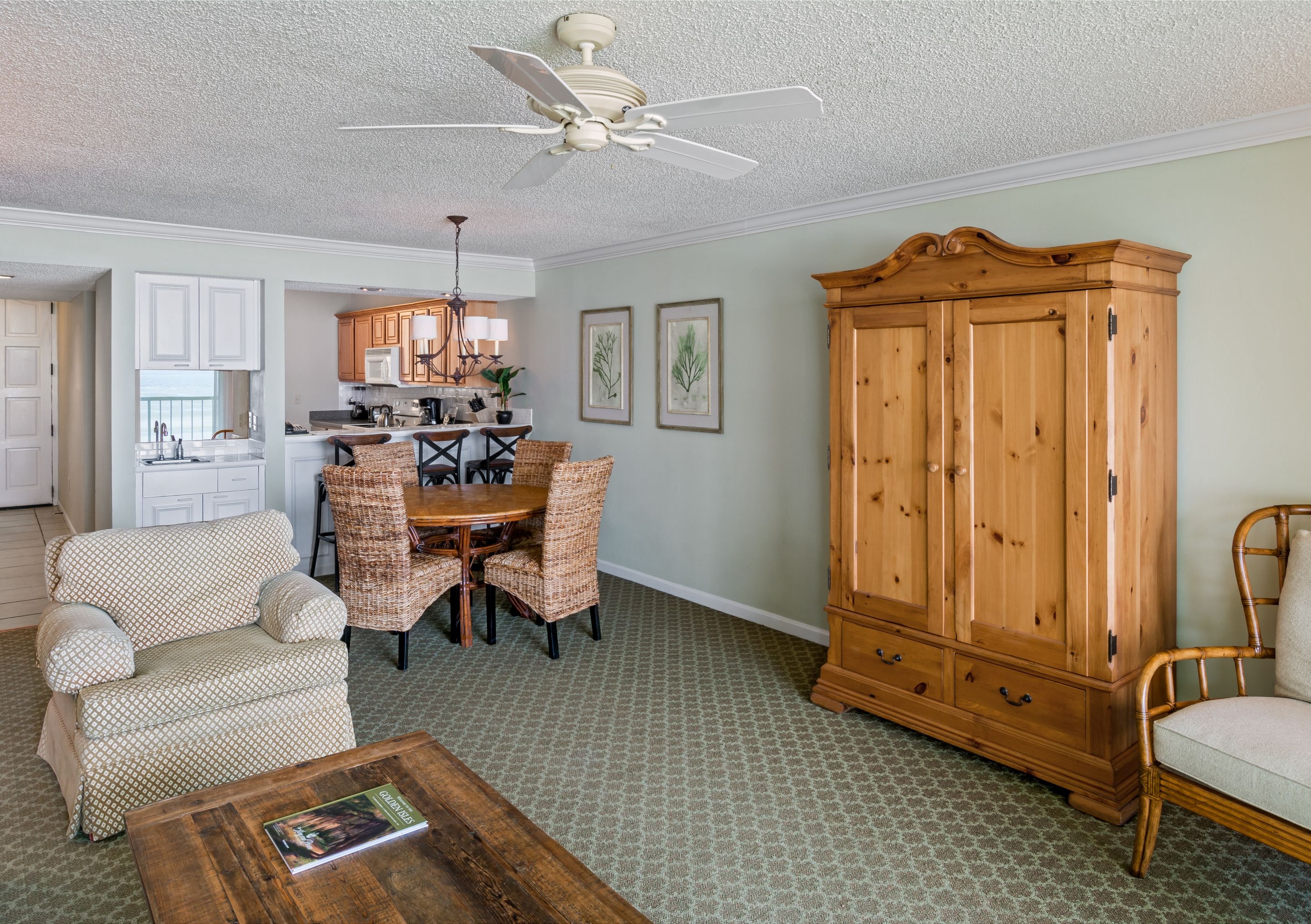 Spacious living and dining area in a coastal suite, featuring patterned green carpet, a large pine armoire, a wooden round dining table with wicker chairs, an upholstered armchair, and a glimpse into a fully equipped kitchen.