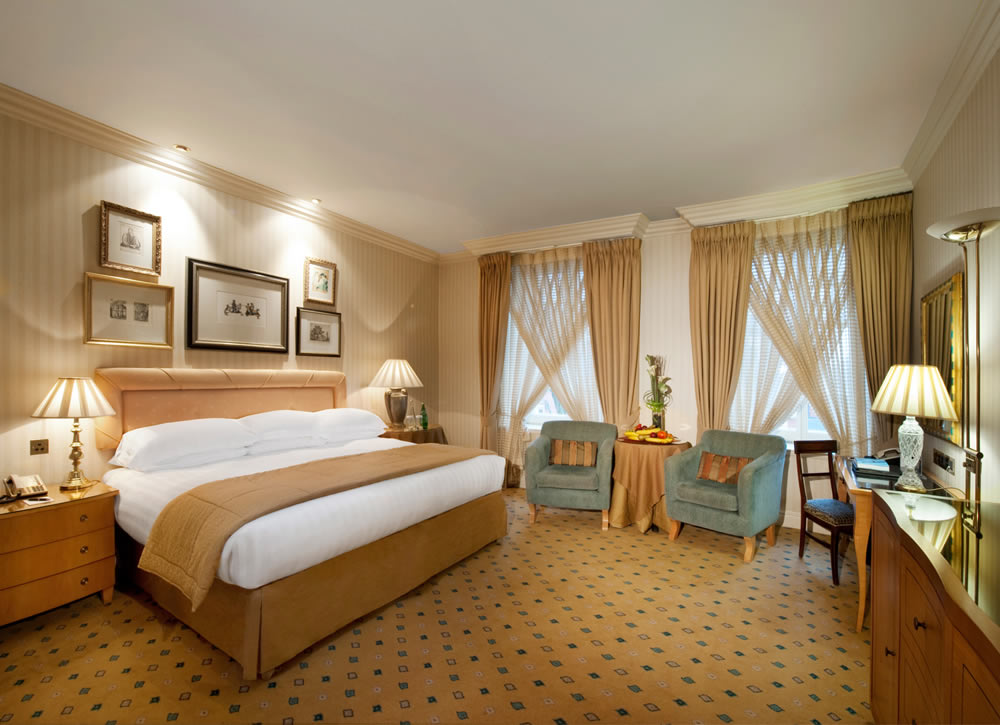 Classic hotel bedroom with beige striped wallpaper, a large bed with a beige runner, framed artwork, and two blue armchairs by draped windows.