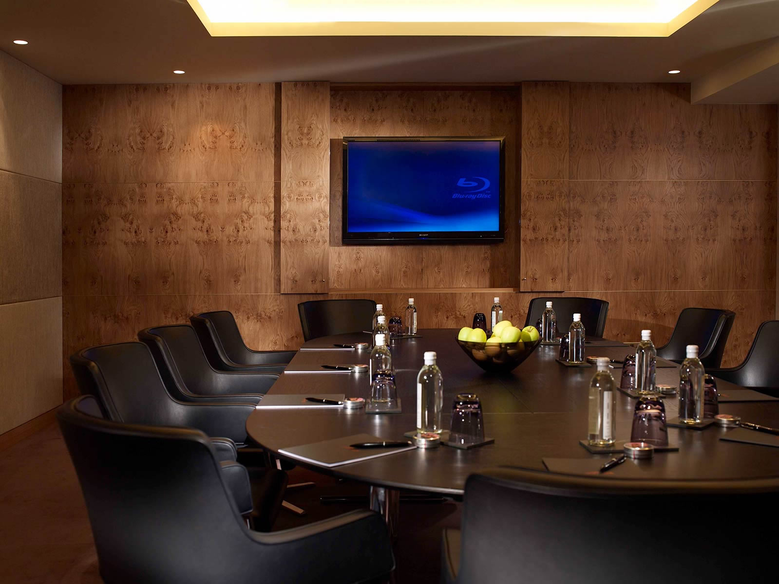 Modern executive boardroom featuring a large oval conference table with black leather armchairs, wood-paneled walls, and a wall-mounted TV screen.