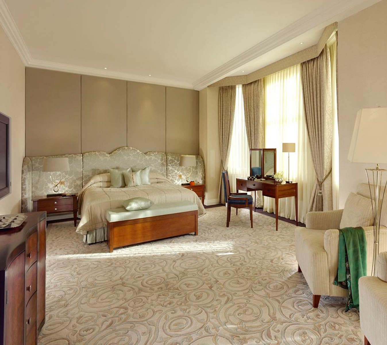 Spacious luxury hotel bedroom in neutral beige tones with a wide patterned headboard, carpet, wooden vanity by tall windows, and an armchair.